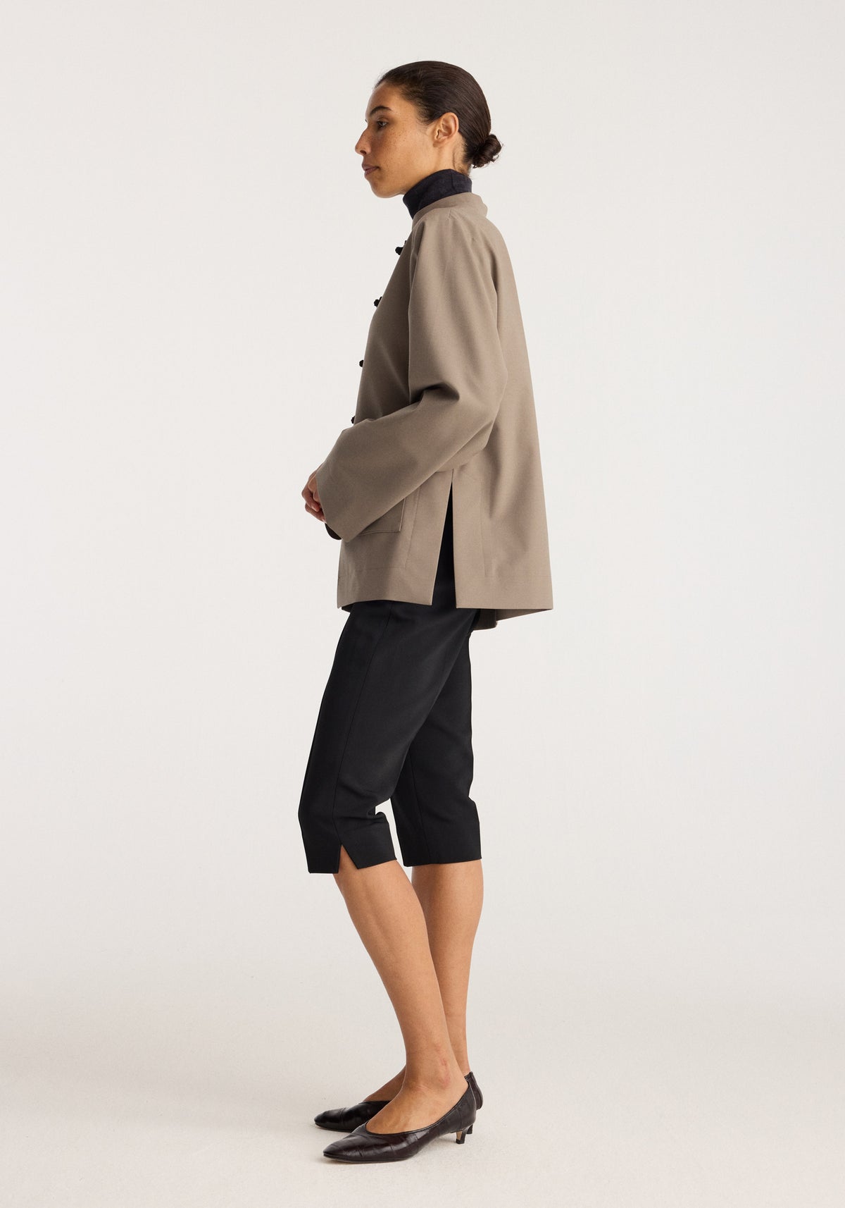 Capri | black worn by a model in profile with a taupe jacket black cropped pants and black pointed heels against a neutral background