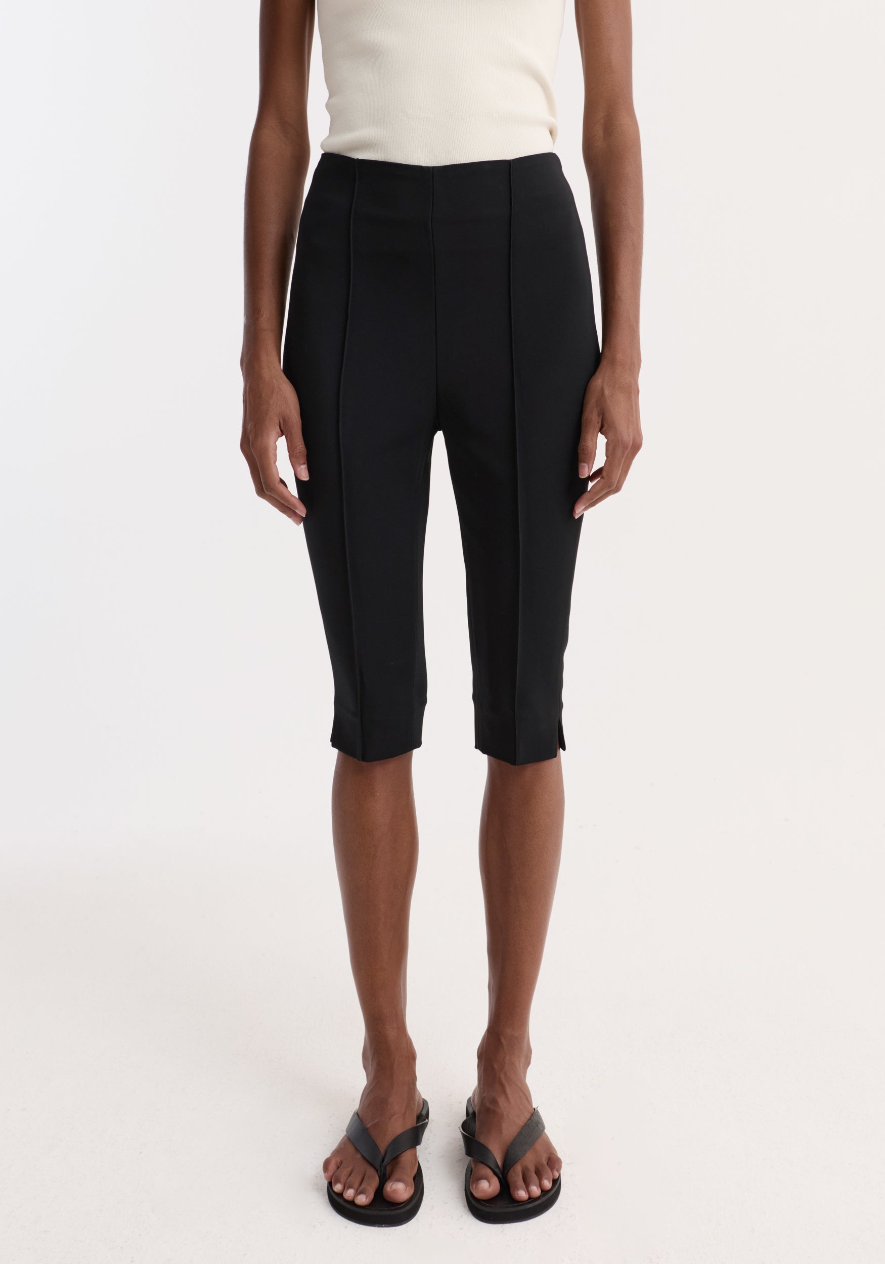 Front view of a person wearing Capri | black fitted knee-length pants with vertical seam details paired with a white sleeveless top and black sandals.