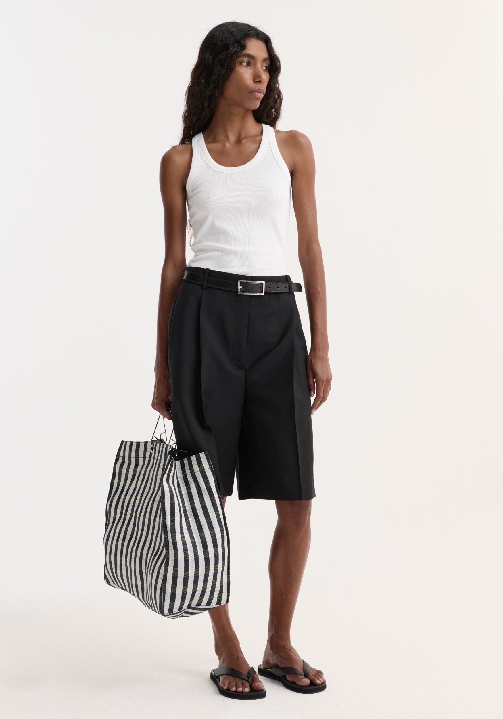 Model wearing Tailored single pleat shorts in black, featuring side pockets and a knee-length cut, paired with a white tank top and striped tote.