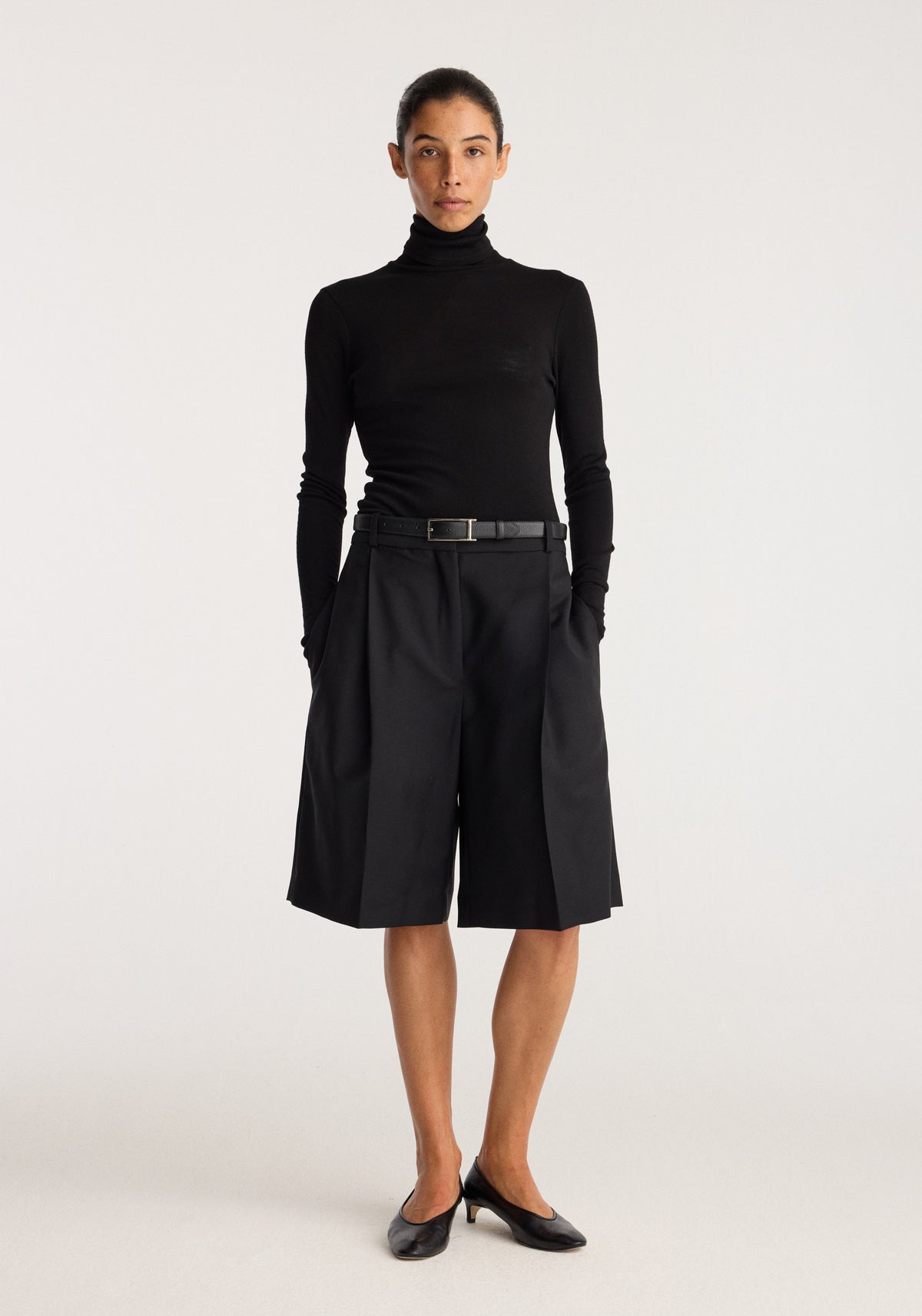 Tailored single pleat shorts | black modeled with a fitted black turtleneck top and belt in a minimalist studio setting