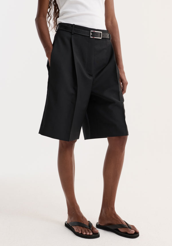 Tailored single pleat shorts | black, featuring side pockets and a knee-length cut, worn with a belt and flip-flops in a studio setting.