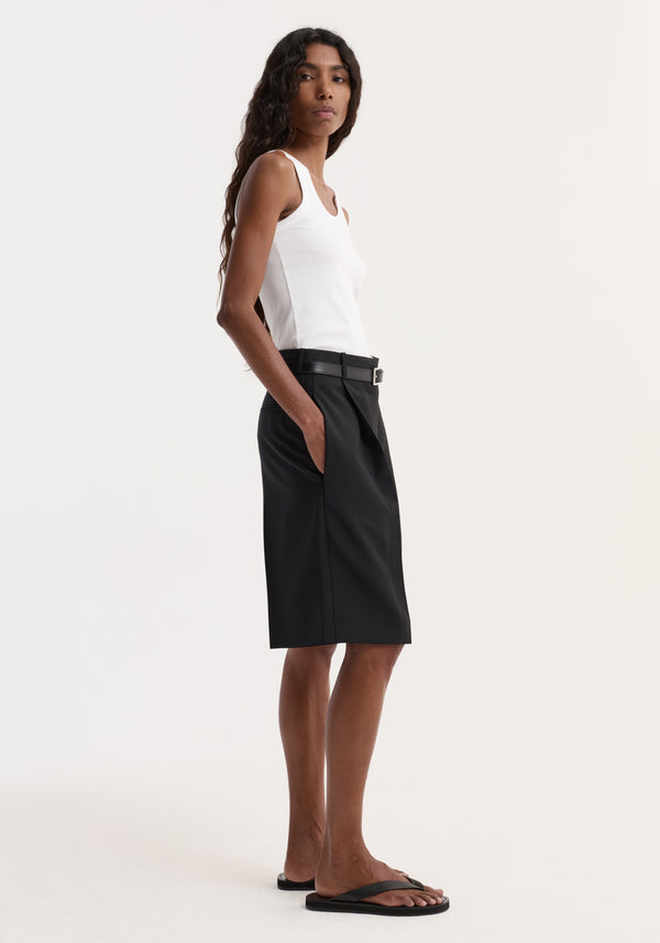Woman modeling black tailored single pleat shorts with side pockets, knee-length fit, and a relaxed posture, wearing black sandals.