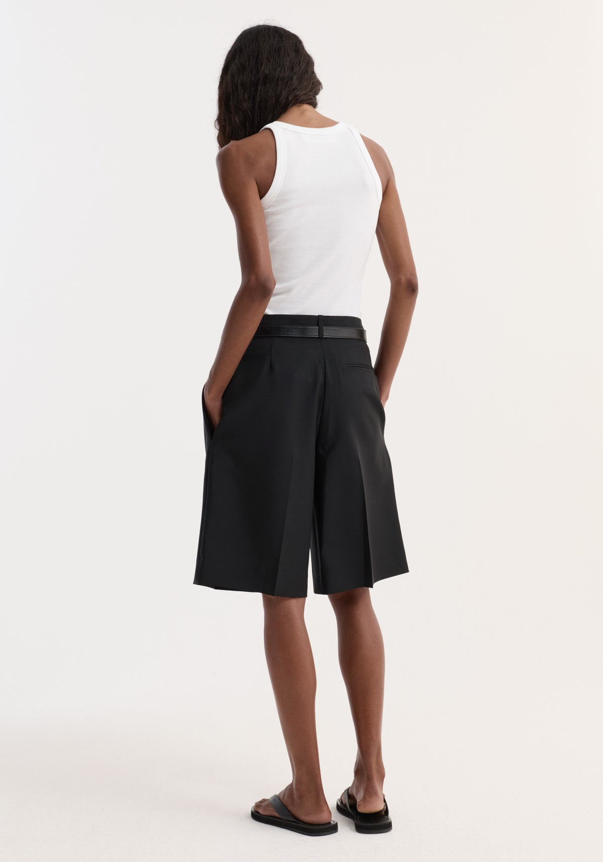 Person wearing Tailored single pleat shorts | black, with side pockets and a slim belt, paired with a white rib-knit tank and black slide sandals.