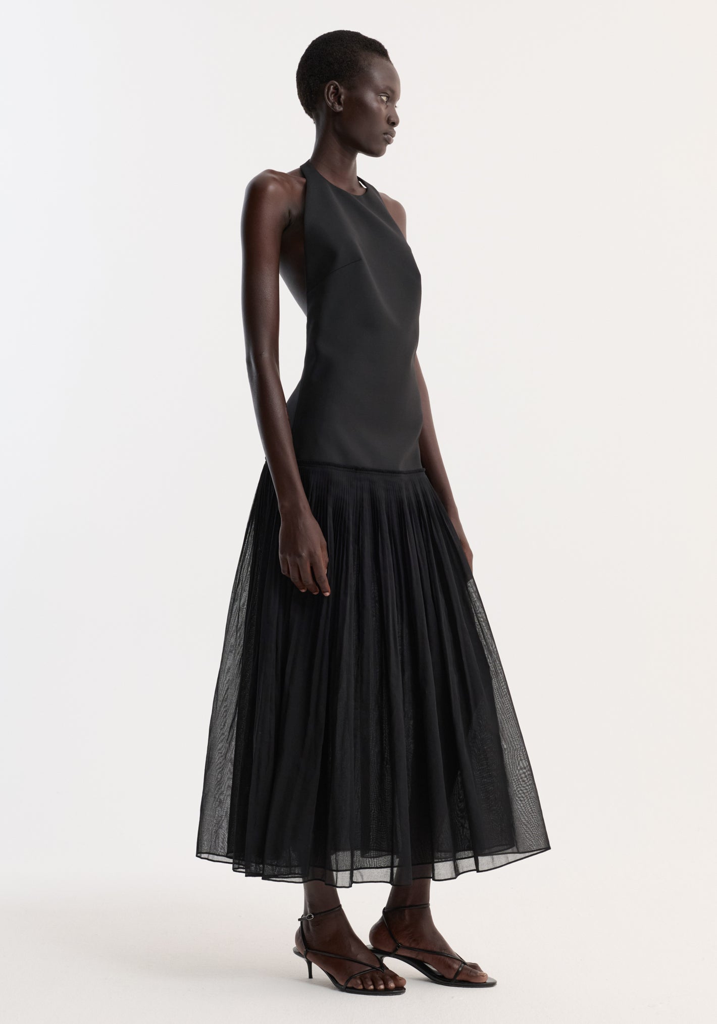 Pleated muslin halter dress | black: Model wears a sleeveless halter top with a structured bodice and a flowing, mid-calf pleated skirt, paired with strappy sandals.