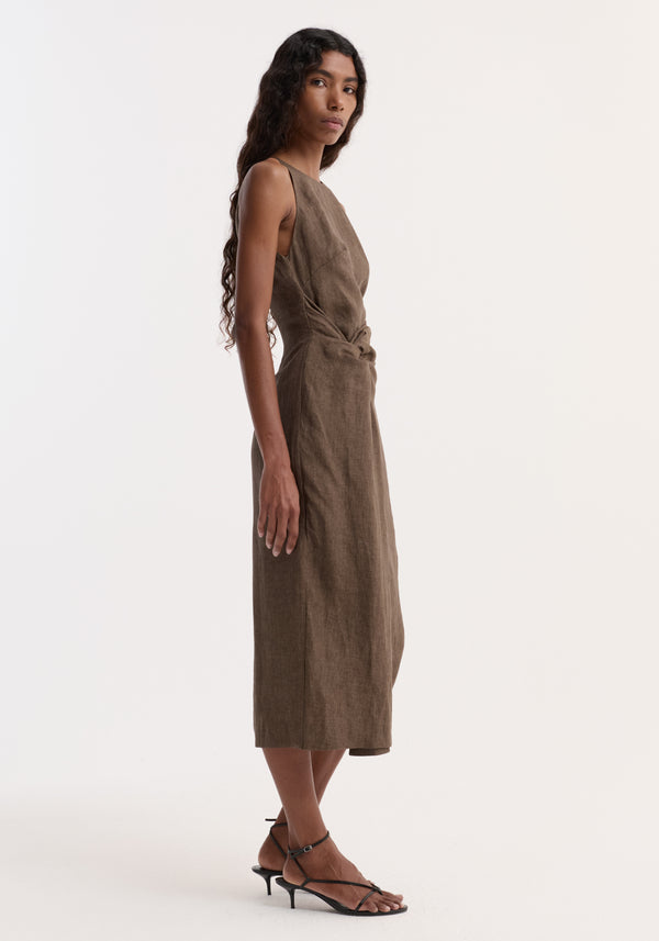 Model in profile wearing Sarong dress | brown melange, showcasing sleeveless, calf-length design with high neckline, sarong-inspired waist knot, and black strappy sandals.