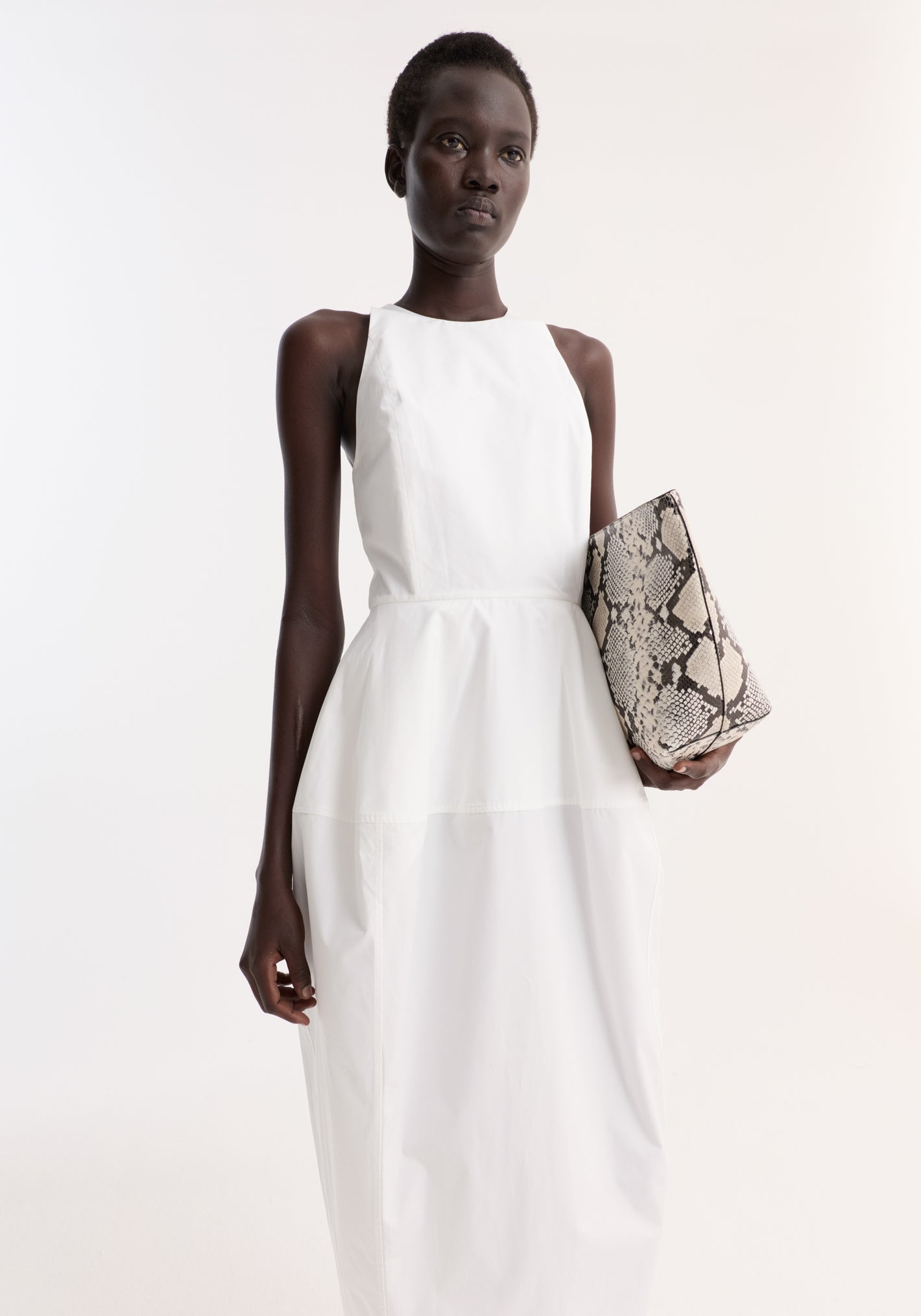 Model wearing Sculptural poplin dress, optic white, with a high neckline and fitted waist, holding a snakeskin-patterned clutch, showcasing its trapezium-shaped skirt and back split.