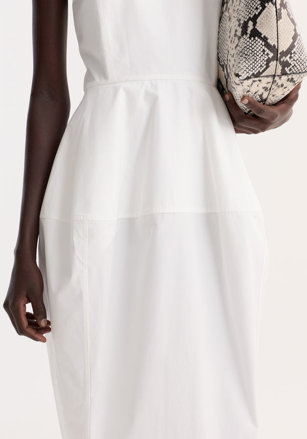 Sculptural poplin dress in optic white, worn by a dark-skinned person, features a tiered waist, trapezium skirt, and back split, paired with snakeskin clutch.