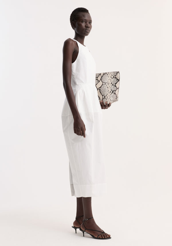 Model wearing Sculptural poplin dress | optic white, featuring a trapezium-shaped skirt and back split, paired with black heeled sandals and snakeskin-print clutch.