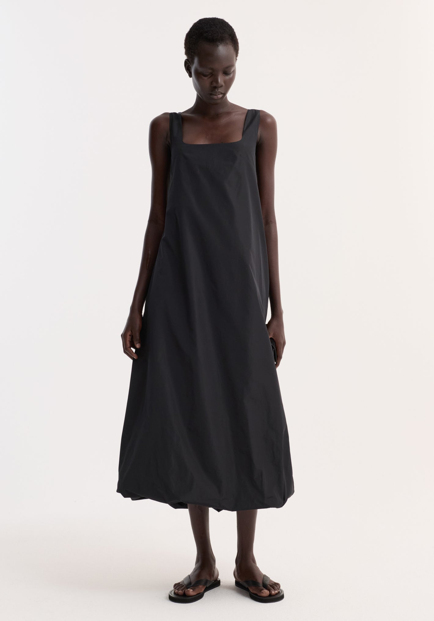 Technical volume dress in black with a sleeveless, square neckline, and balloon hemline, worn by a model standing against a white background.