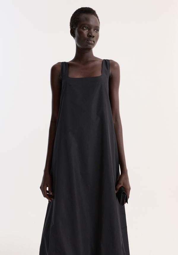 Tall woman in Technical volume dress | black, featuring a sleeveless design, square neckline, balloon hemline, holding a black clutch, showcasing minimalist elegance.
