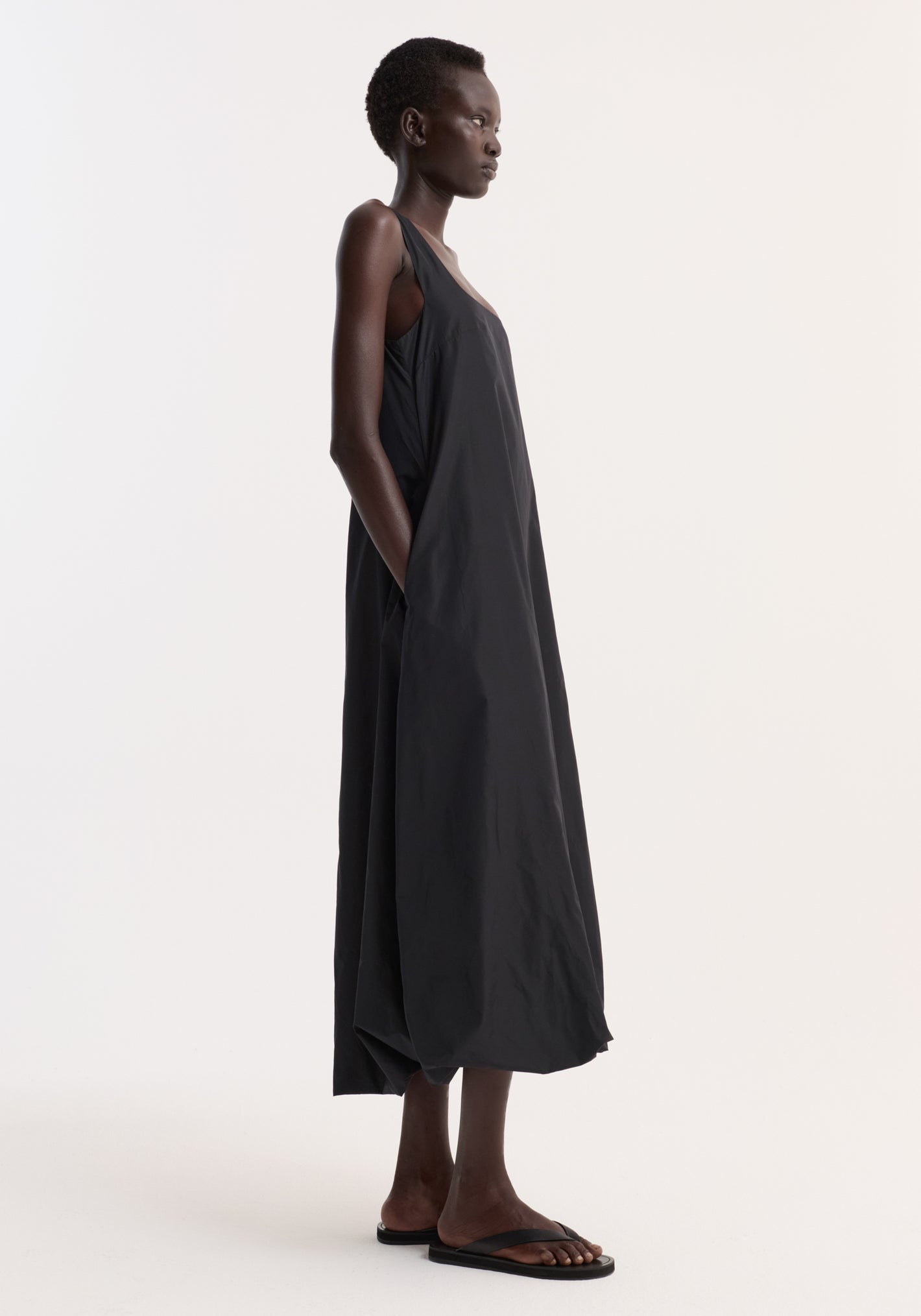 Technical volume dress in black, featuring a sleeveless design with wide straps, a scooped square neckline, and a balloon hemline worn by a model standing in profile.