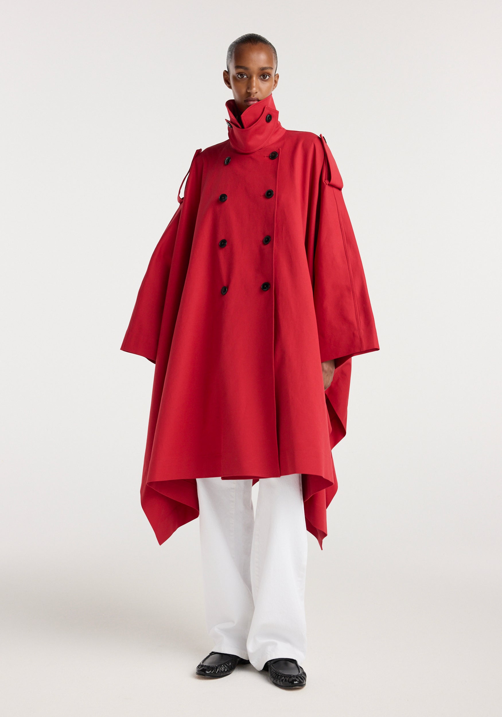 Model wearing a bright red Cape trenchcoat | cherry with a high collar, double-breasted black buttons, wide sleeves, and an asymmetrical hem, paired with white pants and black shoes.