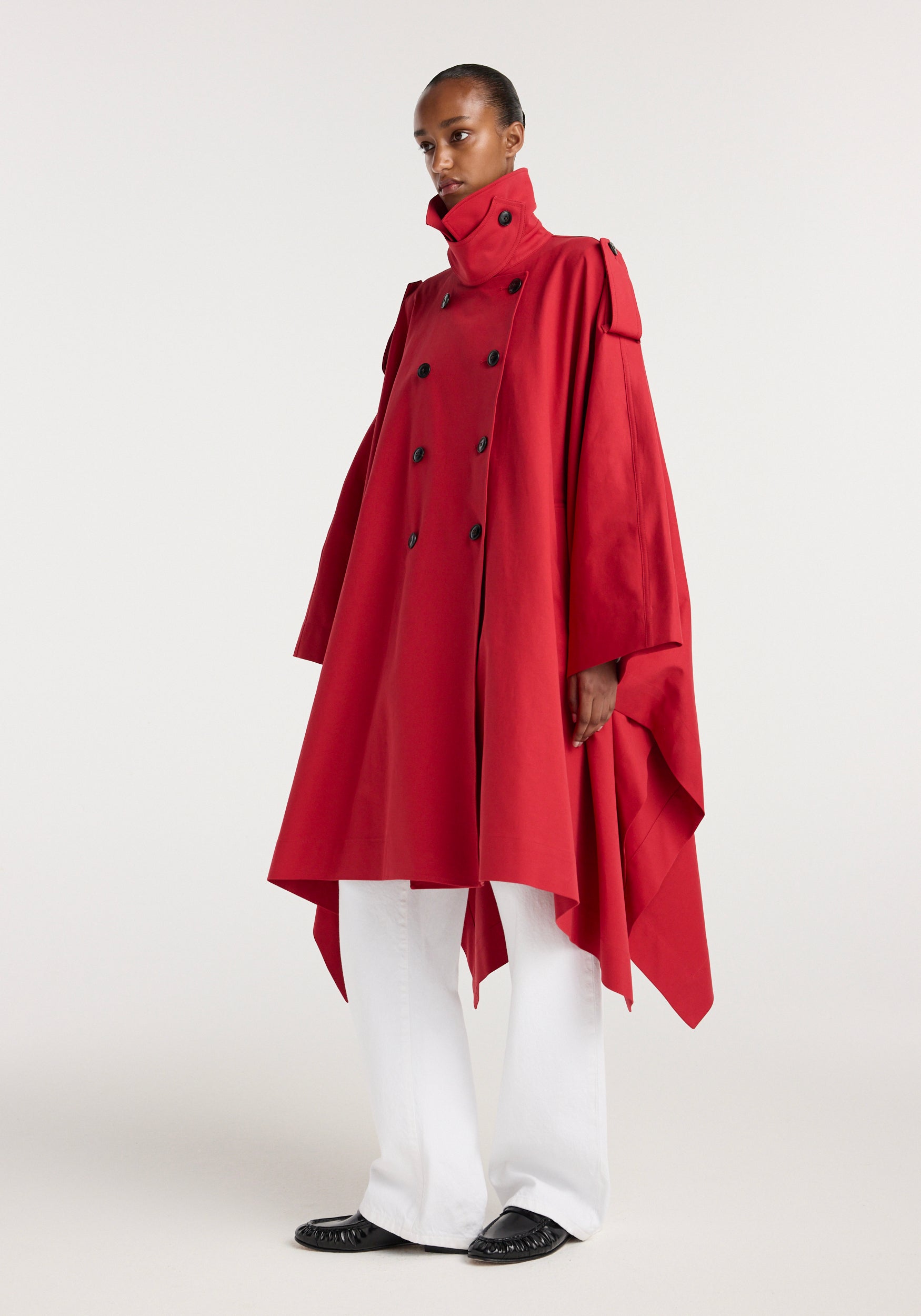 Model wearing a flowing red cape trenchcoat | cherry with a high collar, double-breasted buttons, and asymmetrical hem paired with white pants and black shoes against a plain background.