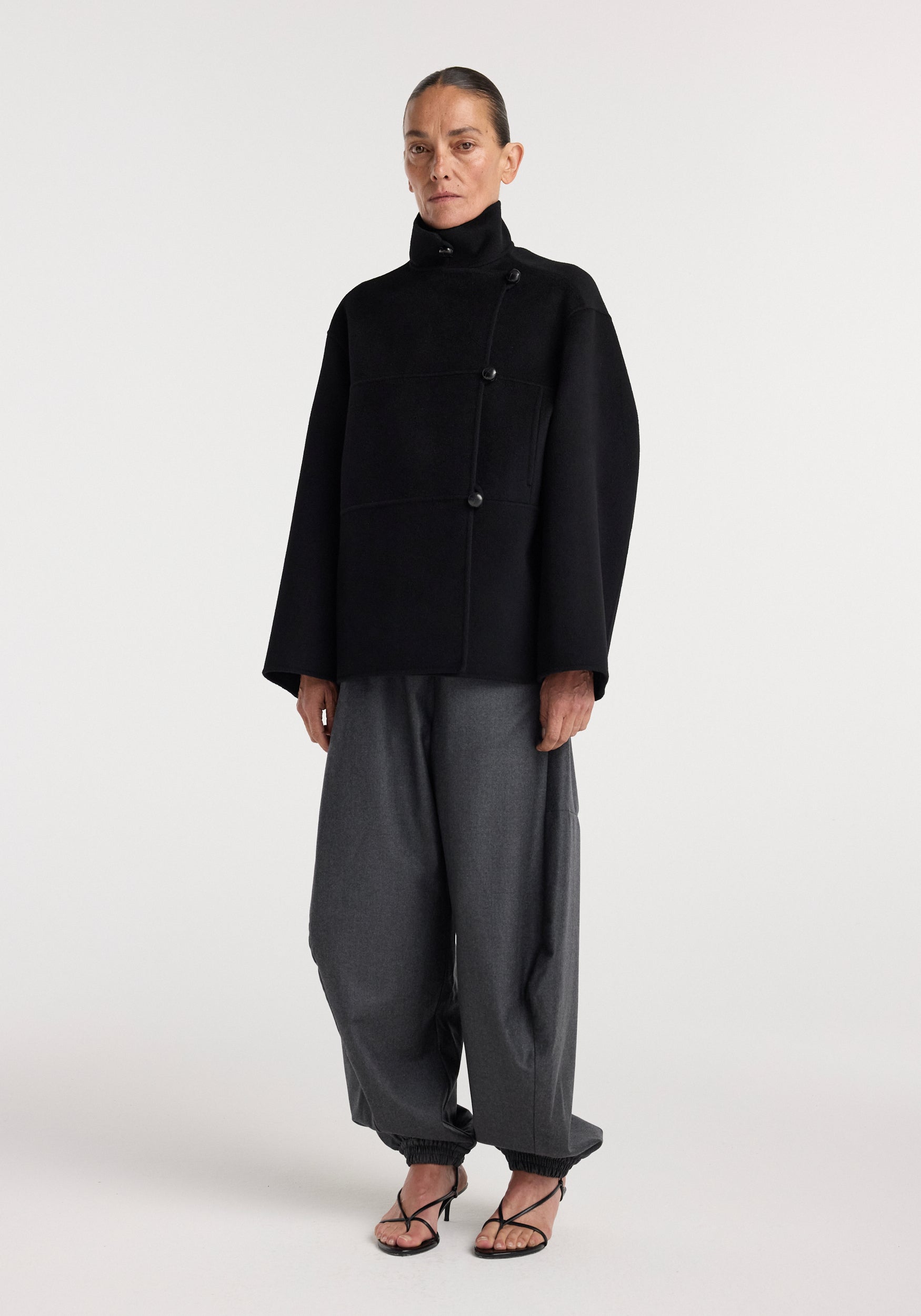 Woman wearing a black double-faced wool jacket with an asymmetrical button closure, paired with loose gray pants and black strappy heels, standing against a plain background.