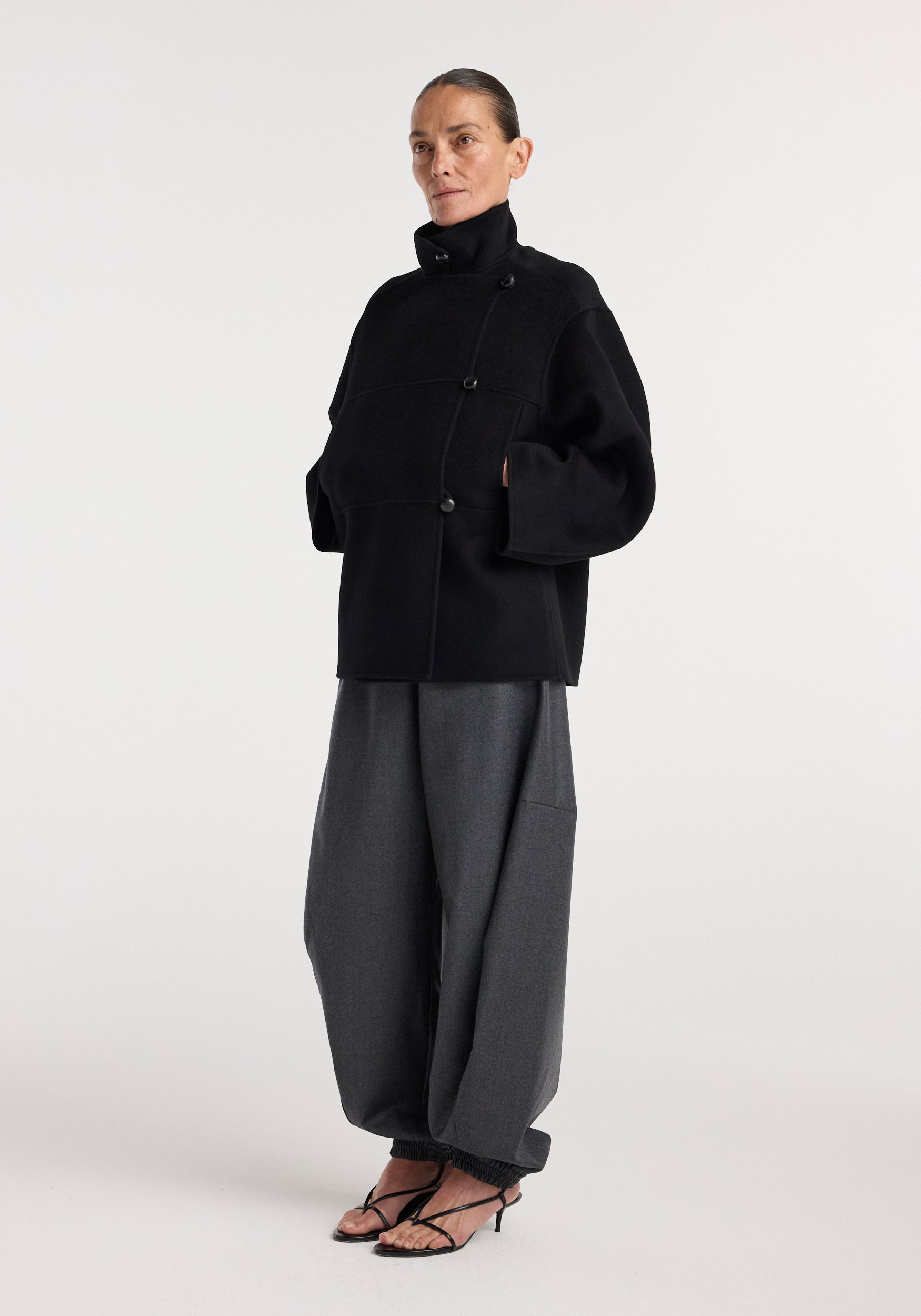Woman wearing a black Double-faced wool jacket | black with an asymmetrical button design, paired with wide-leg gray trousers and black heeled sandals, standing against a plain background.