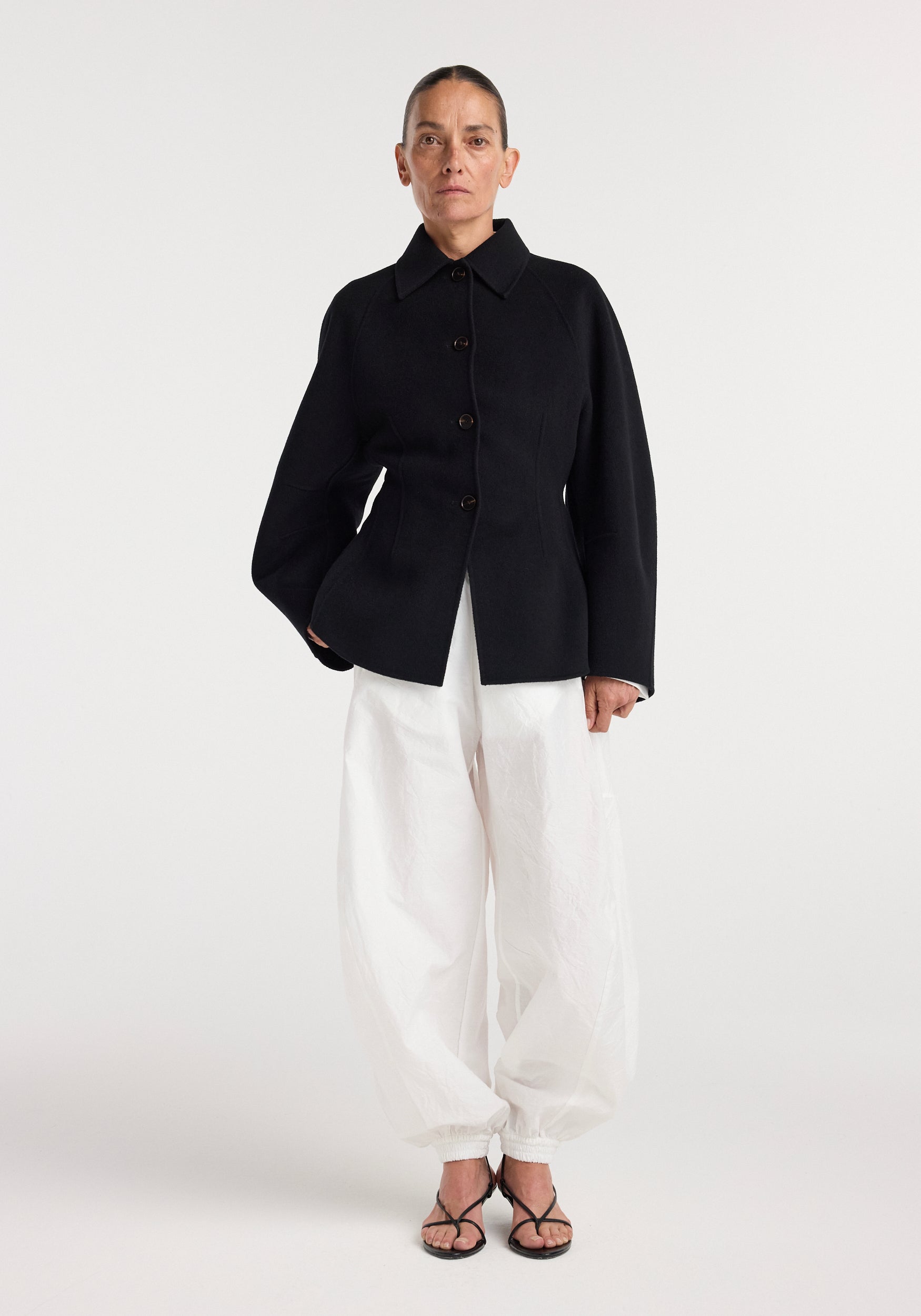 Woman wearing a minimalist black double-faced sculptural jacket | black with large buttons, paired with loose white pants and black strappy heels, standing against a white background.