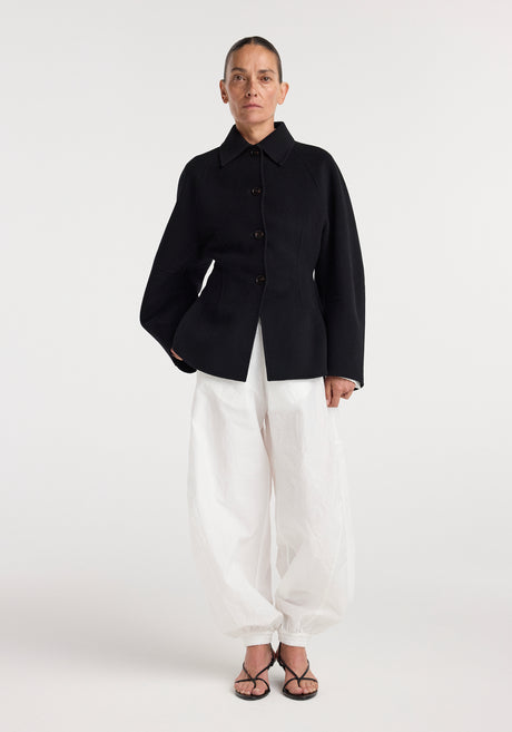Woman wearing a minimalist black double-faced sculptural jacket | black with large buttons, paired with loose white pants and black strappy heels, standing against a white background.