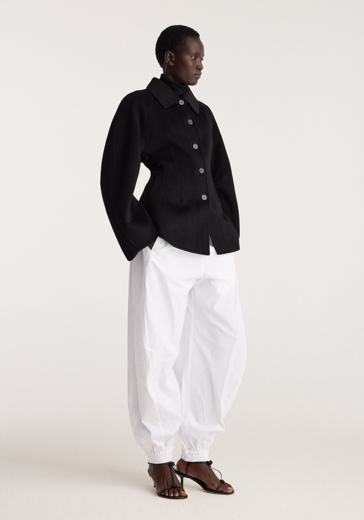 Double-faced sculptural jacket | black worn with white trousers and black strappy sandals in a minimalist studio setting
