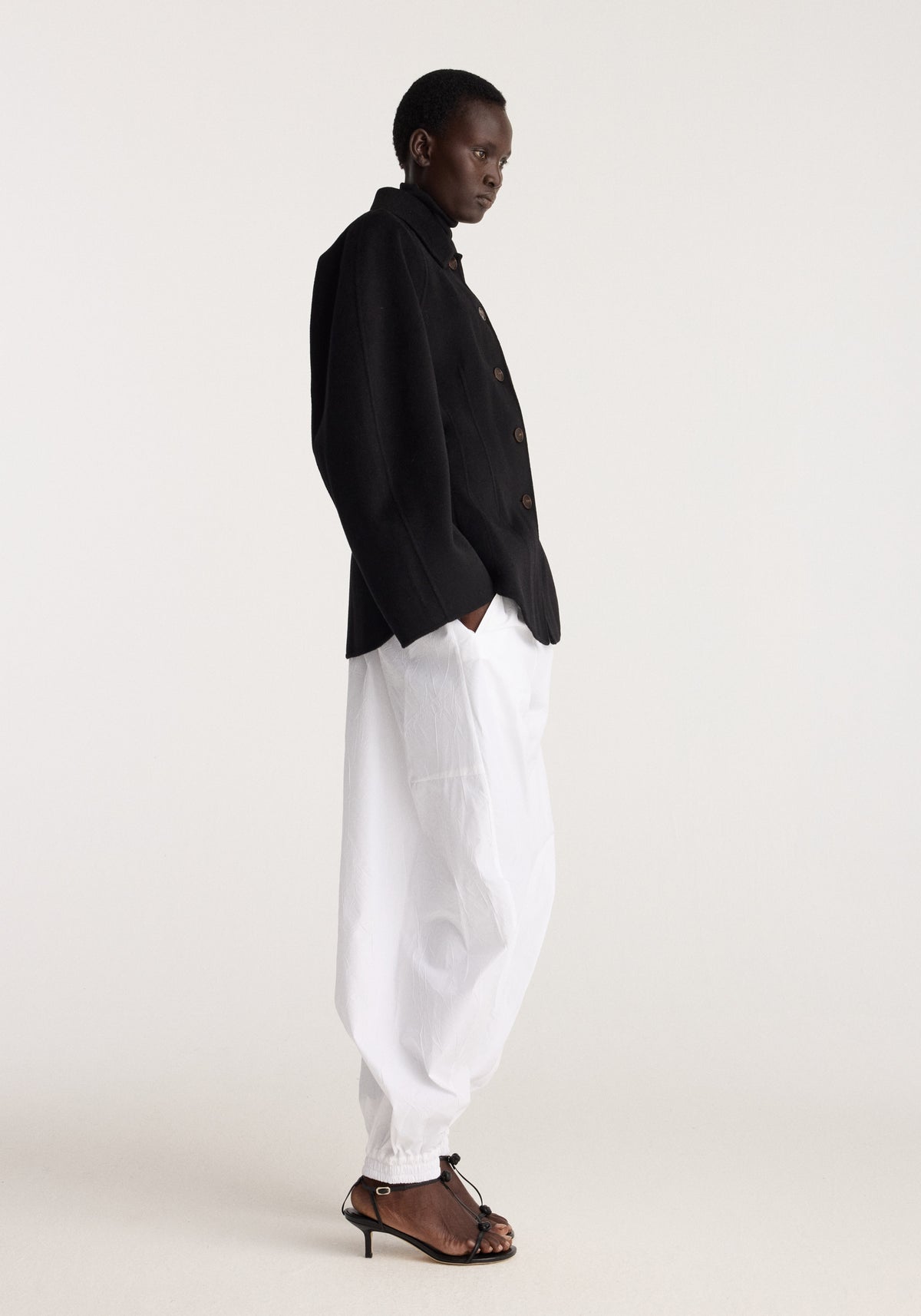 Double-faced sculptural jacket | black worn by a model with white trousers and brown sandals in a minimalist studio setting