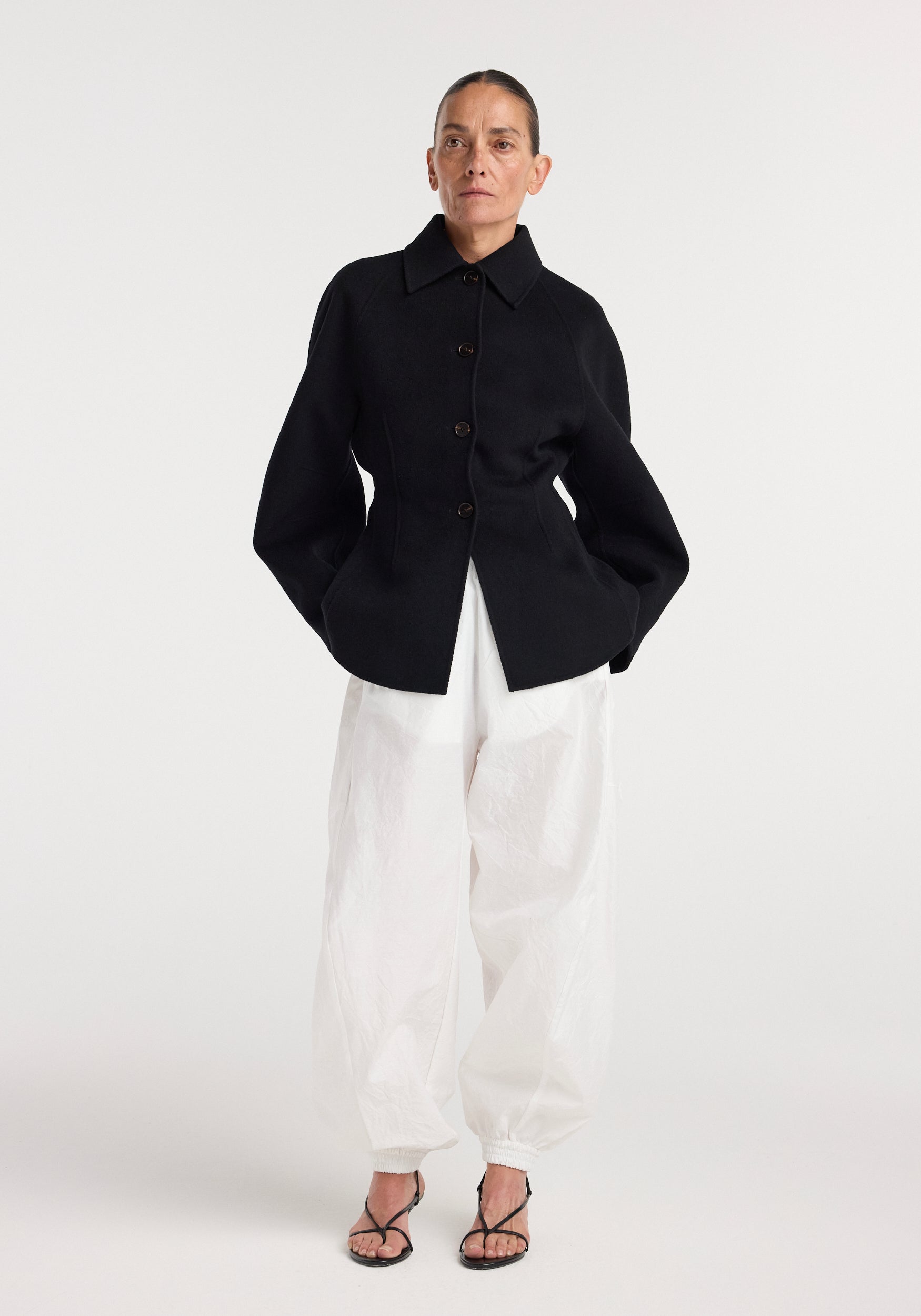 Model wearing a structured black button-up double-faced sculptural jacket | black paired with loose white pants and black strappy heels against a plain background.