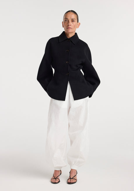 Woman wearing a minimalist black double-faced sculptural jacket | black with large buttons, paired with loose white pants and black strappy heels, standing against a white background.