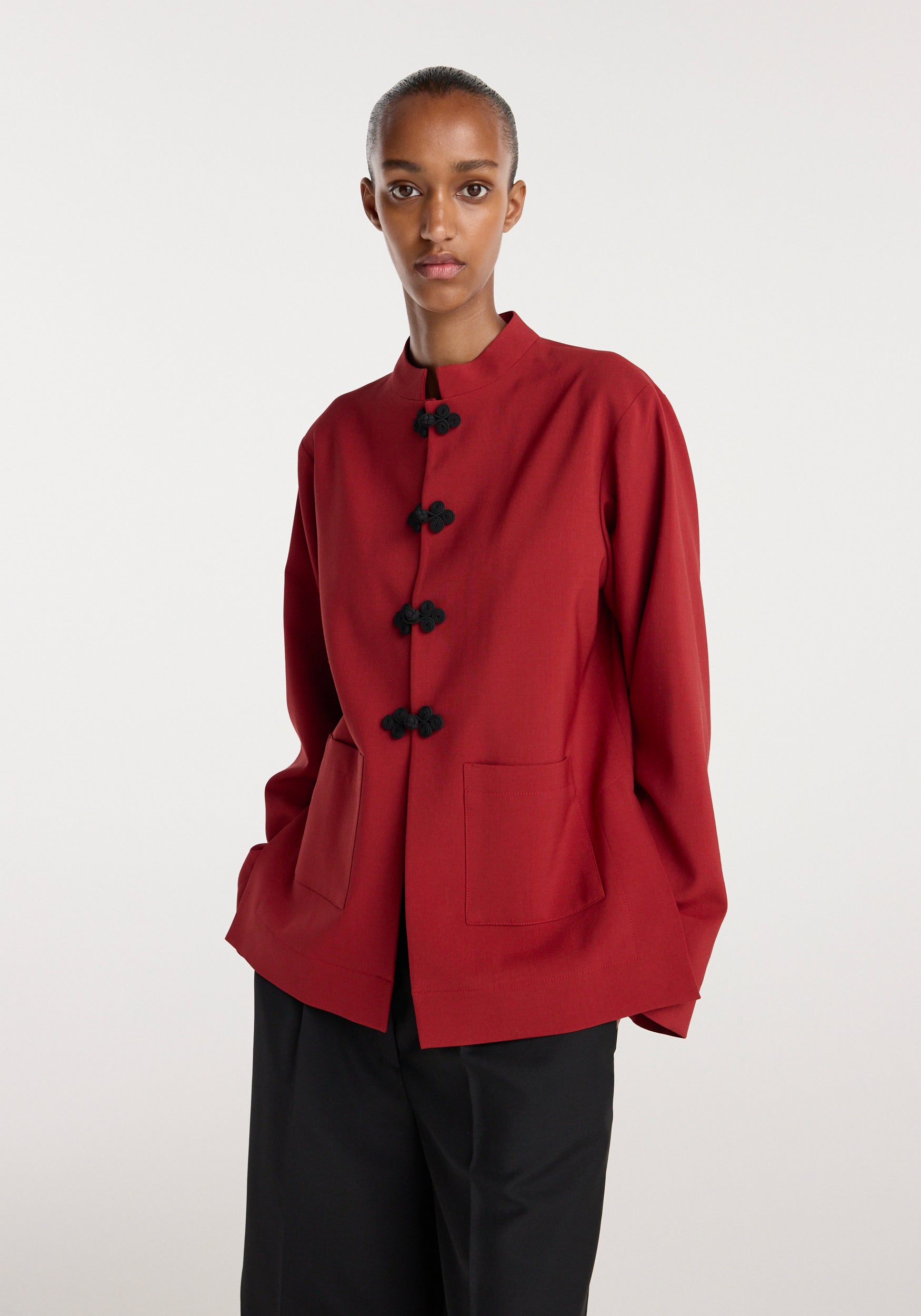 Model wearing a carmine red jacket with traditional black pankou closures and patch pockets, paired with loose black pants, against a plain white background, Pankou closure jacket | carmine.