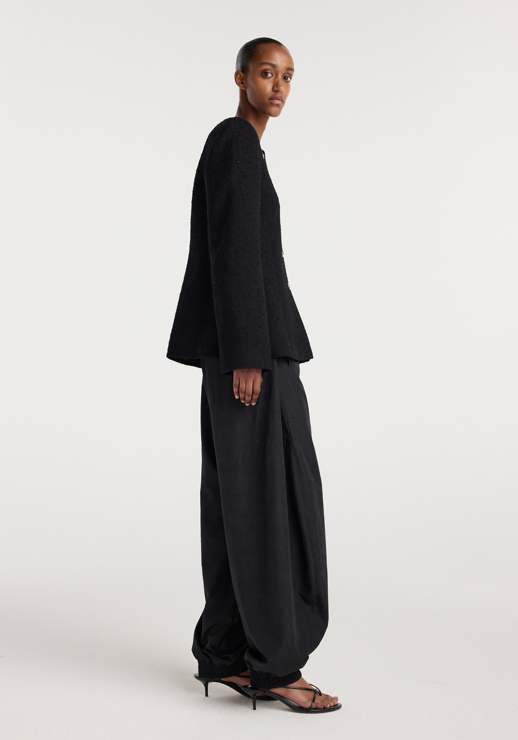 Side view of a model wearing a black Bouclé shaped blazer | black paired with loose black pants and black open-toe heels against a plain white background.