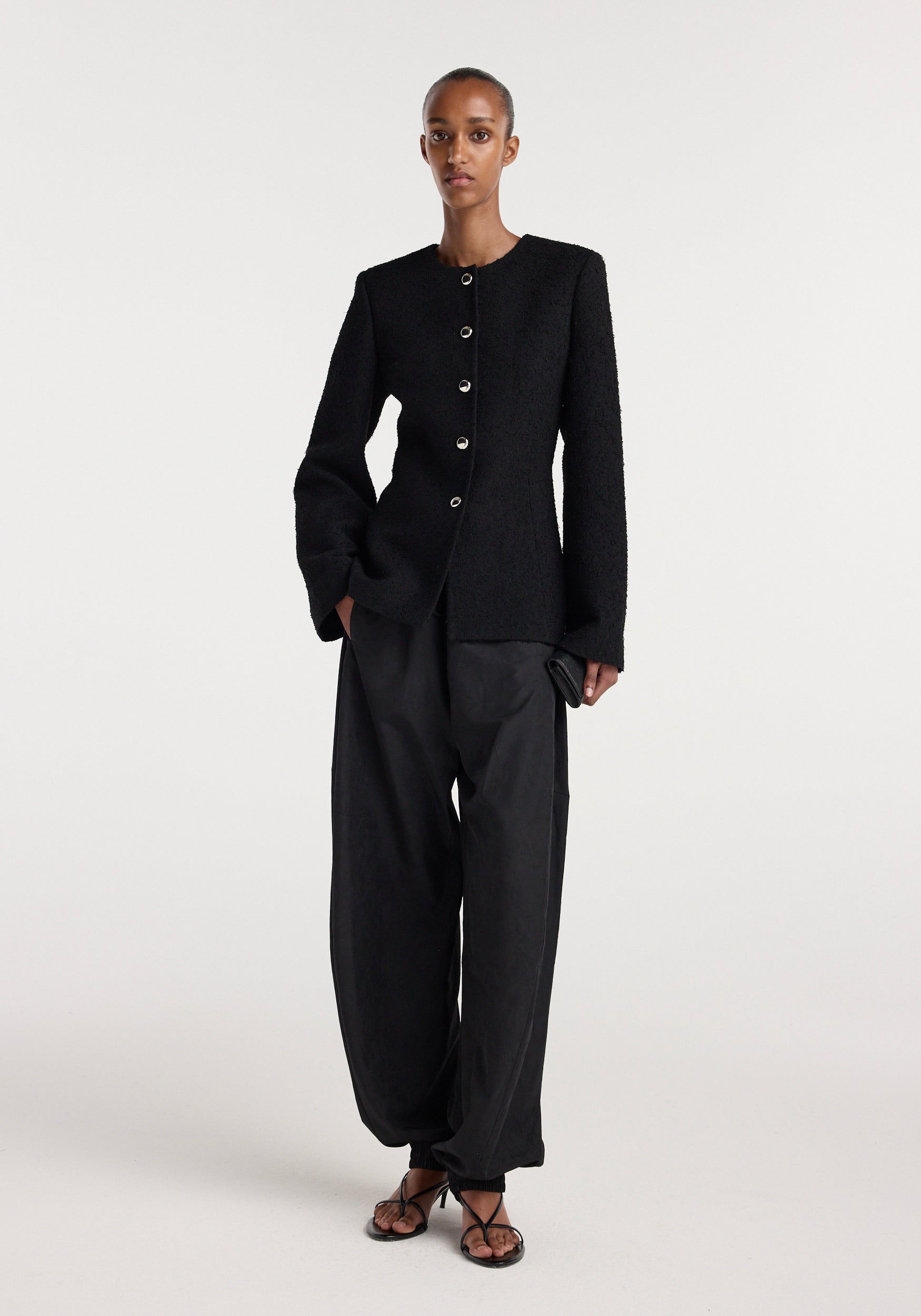 Model wearing a black bouclé shaped blazer | black with a round neckline and silver buttons, paired with loose black pants and black strappy sandals on a plain background.