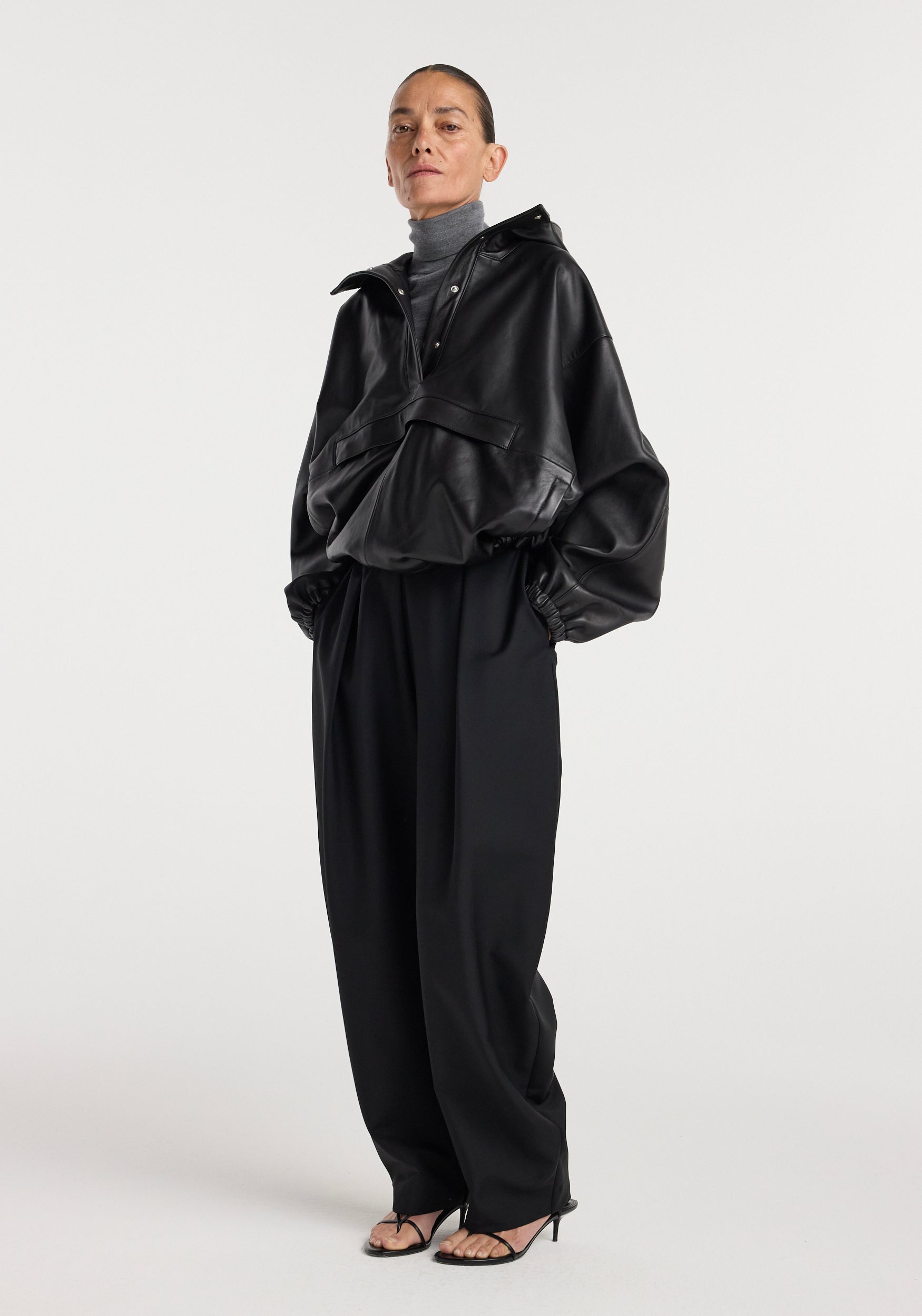 Woman wearing a black leather oversized anorak | black with a high collar and front pockets, styled with a gray turtleneck, wide-leg black pants, and black high-heeled sandals.