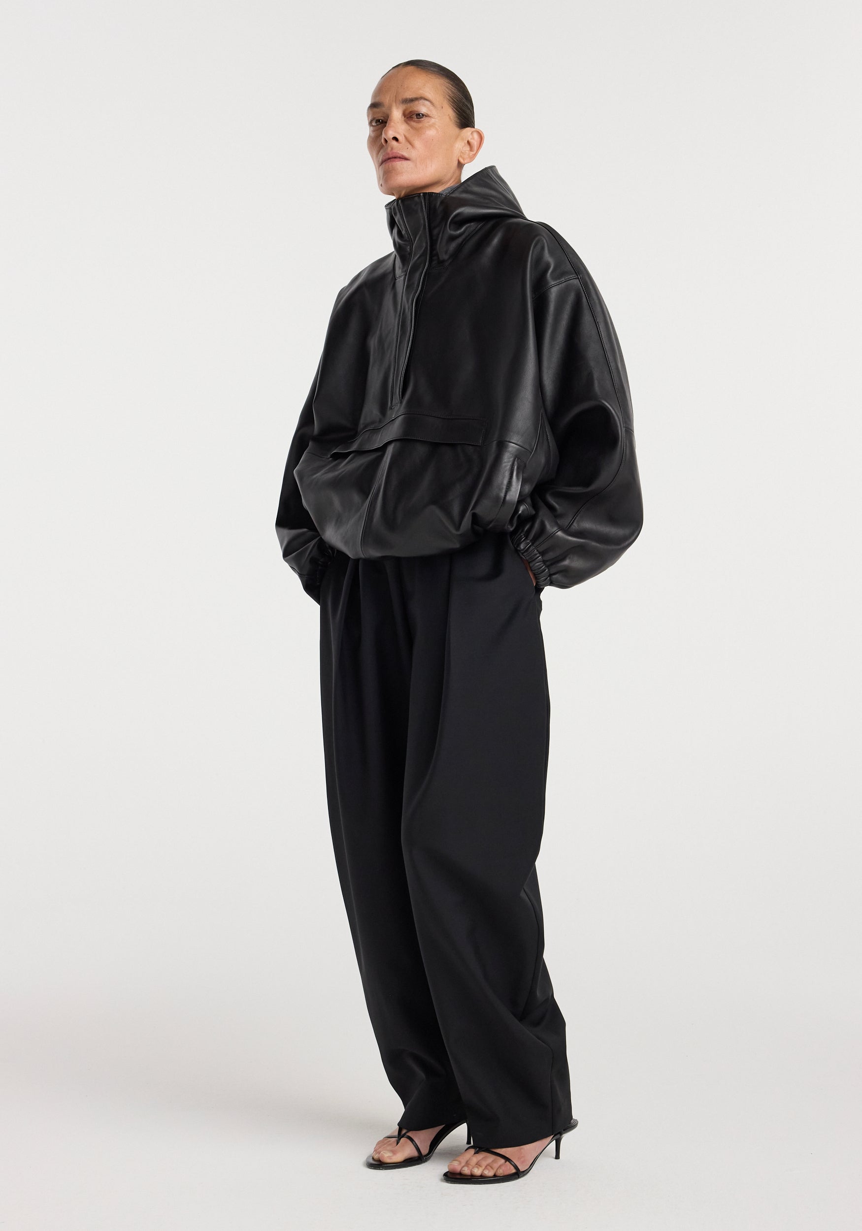 A model wearing a black Leather oversized anorak | black with a high collar and front pocket, paired with wide-leg black pants and black heeled sandals against a plain background.