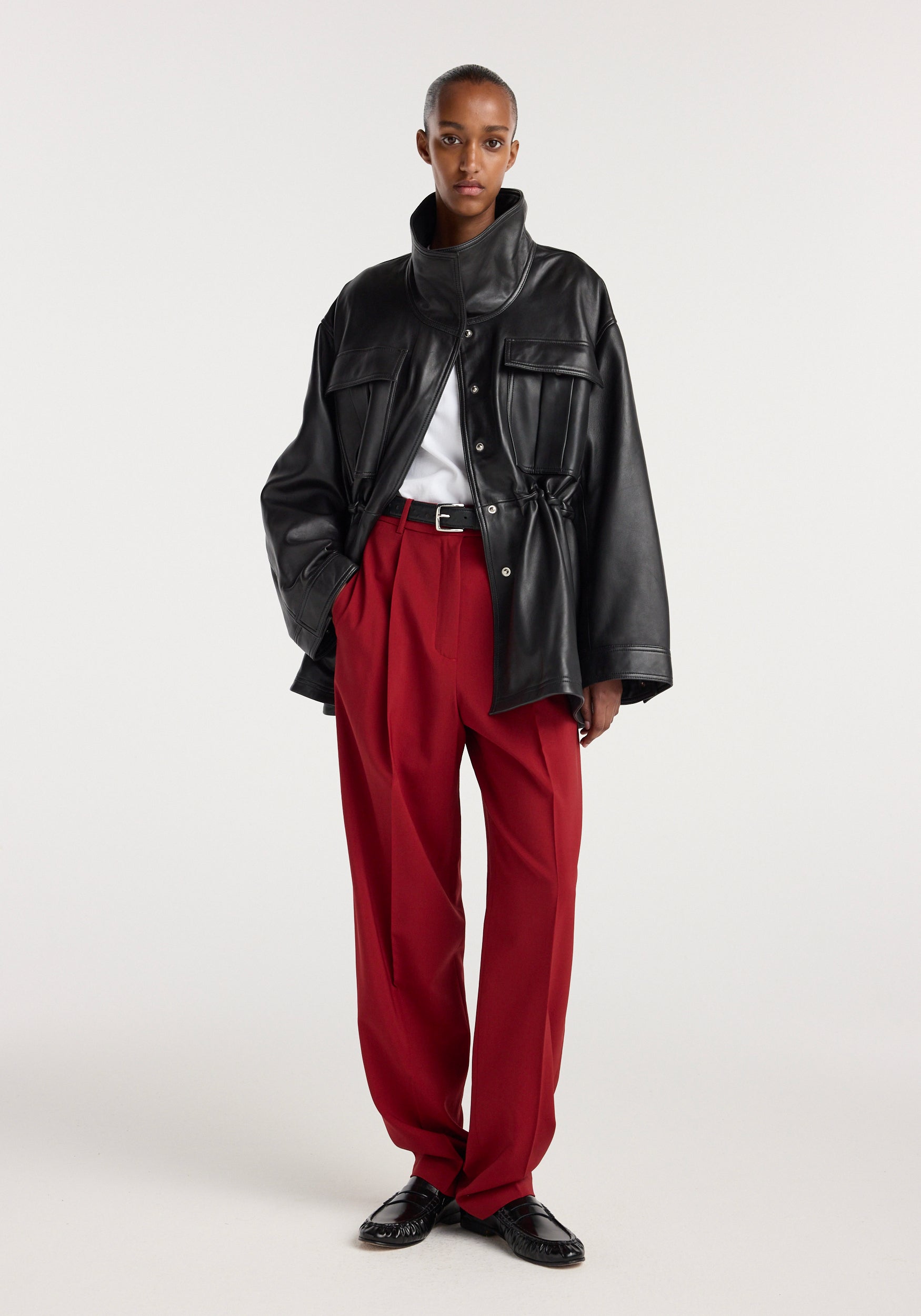 Model wearing a black oversized sculptural leather jacket | black with high collar and snap buttons, paired with red tailored trousers, a white top, and black shoes.
