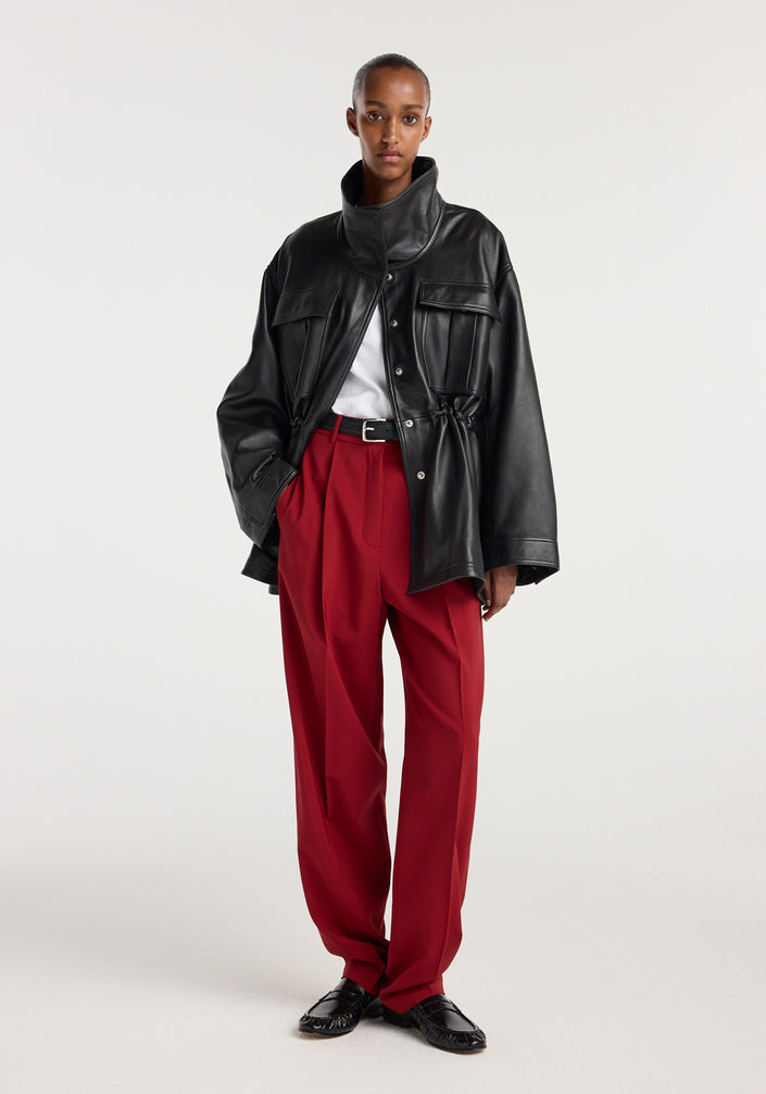 Model wearing a black oversized sculptural leather jacket | black with high collar and snap buttons, paired with red tailored trousers, a white top, and black shoes.