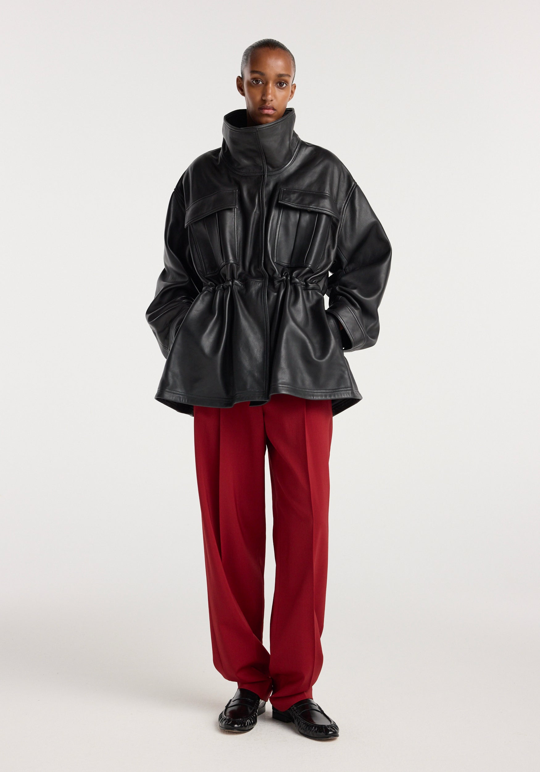 Model wearing a black sculptural leather jacket with a high collar and cinched waist paired with loose red trousers and black shoes, standing against a plain background.