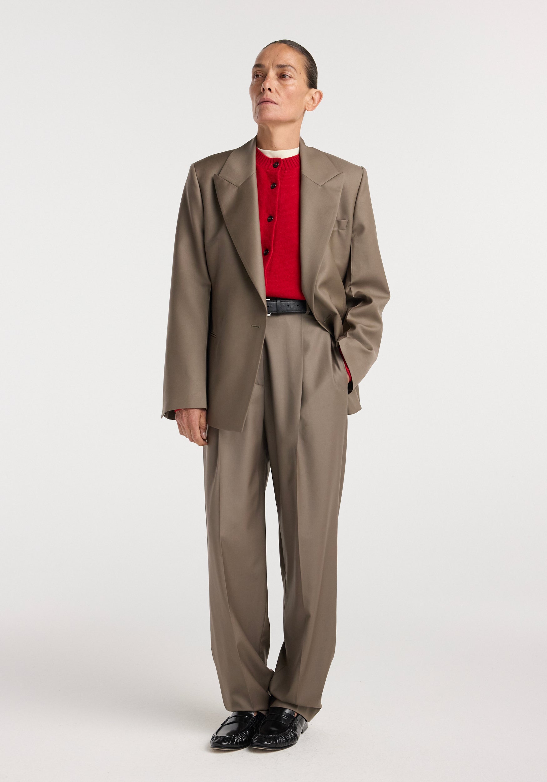 Model wearing an hourglass shaped blazer | desert brown paired with matching trousers, black belt, red buttoned shirt, and black shoes against a plain white background.