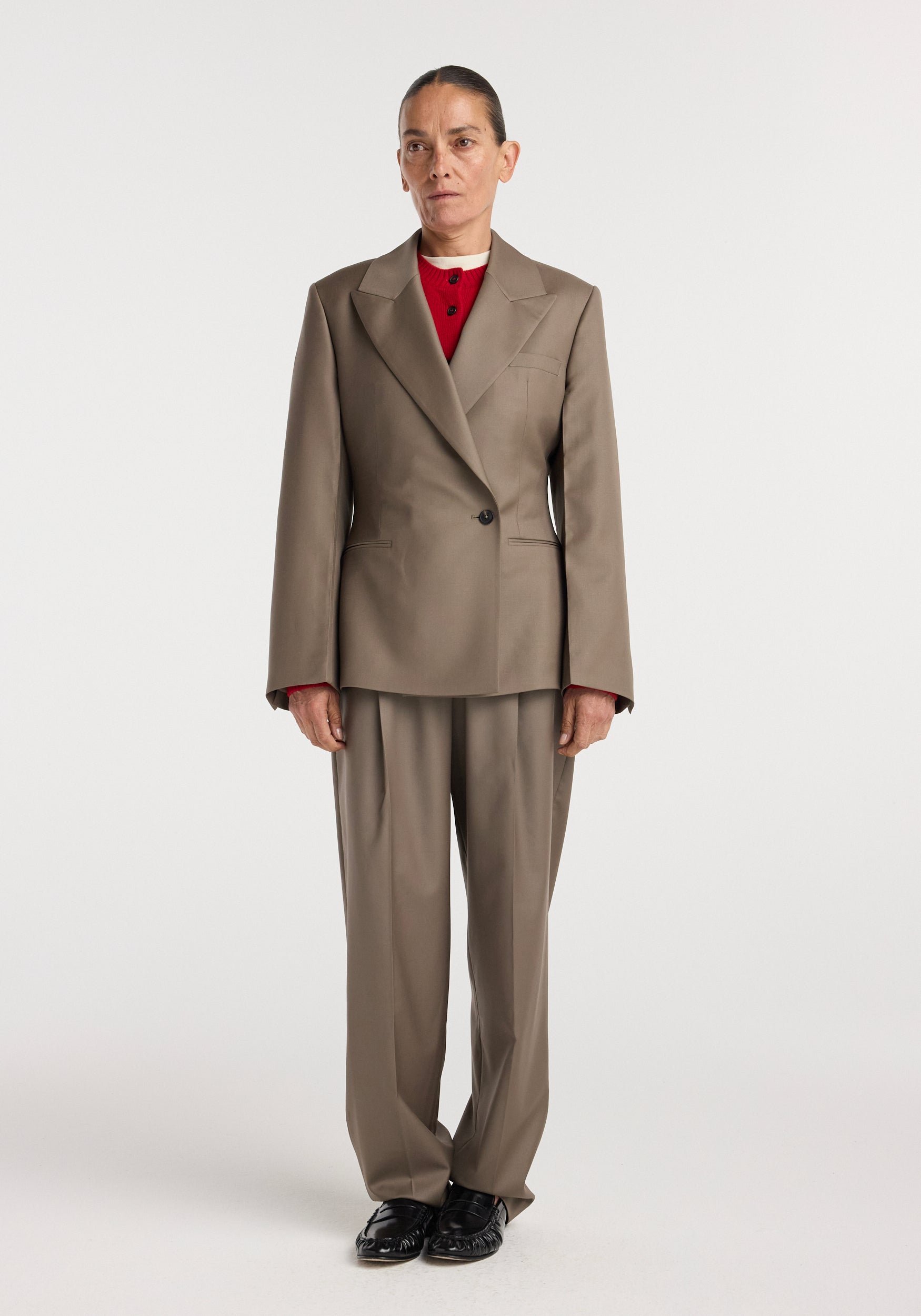 A woman wearing an hourglass shaped blazer | desert brown paired with matching wide-leg trousers, a red top underneath, and black shoes, standing against a plain white background.