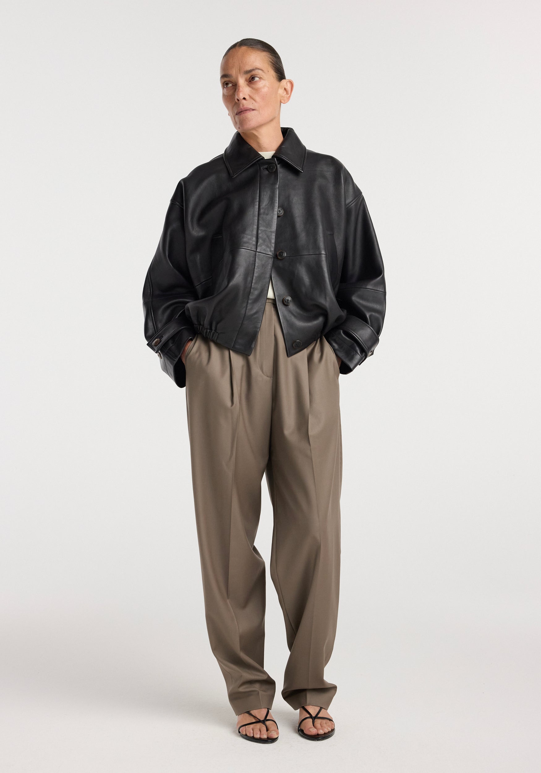 Woman wearing a black Leather blouson jacket | black with loose khaki pants and black strappy sandals, posing against a plain white background.
