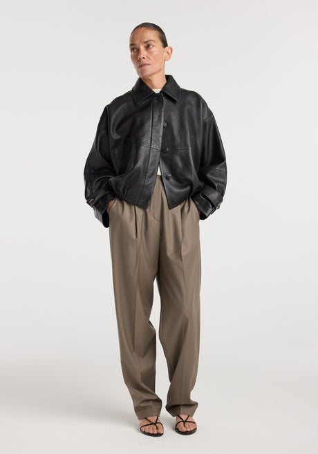 Woman wearing a black Leather blouson jacket | black with loose khaki pants and black strappy sandals, posing against a plain white background.