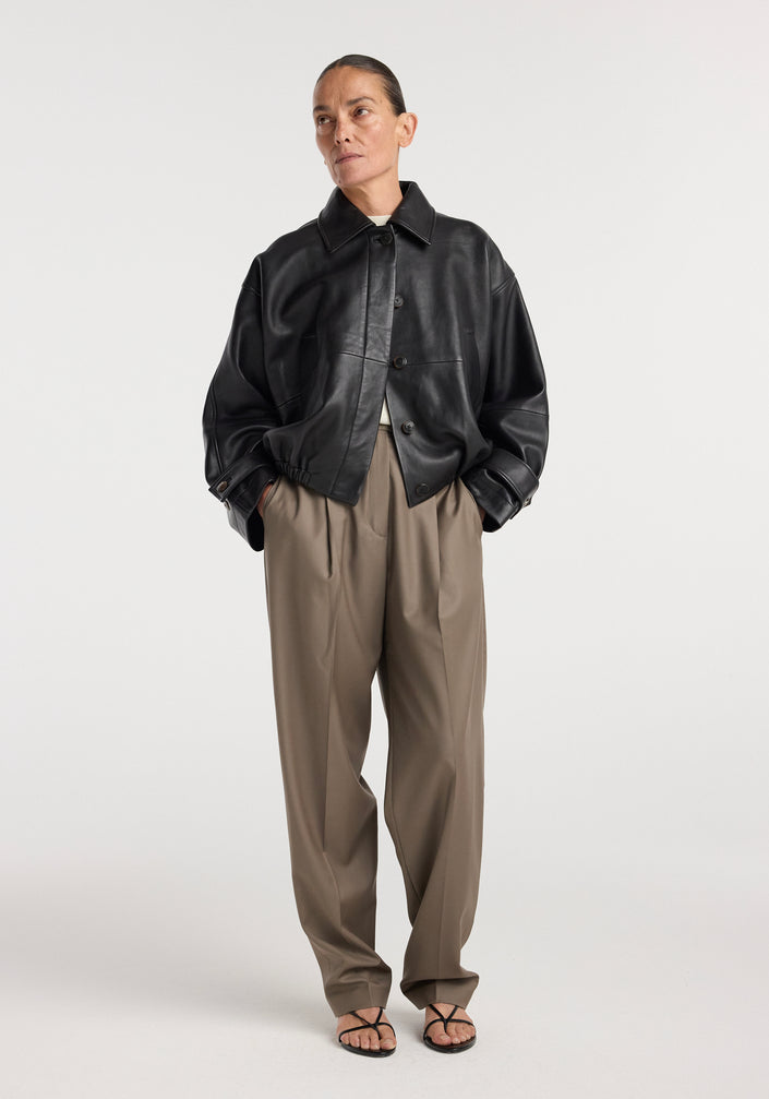 Woman wearing a black Leather blouson jacket | black with loose khaki pants and black strappy sandals, posing against a plain white background.