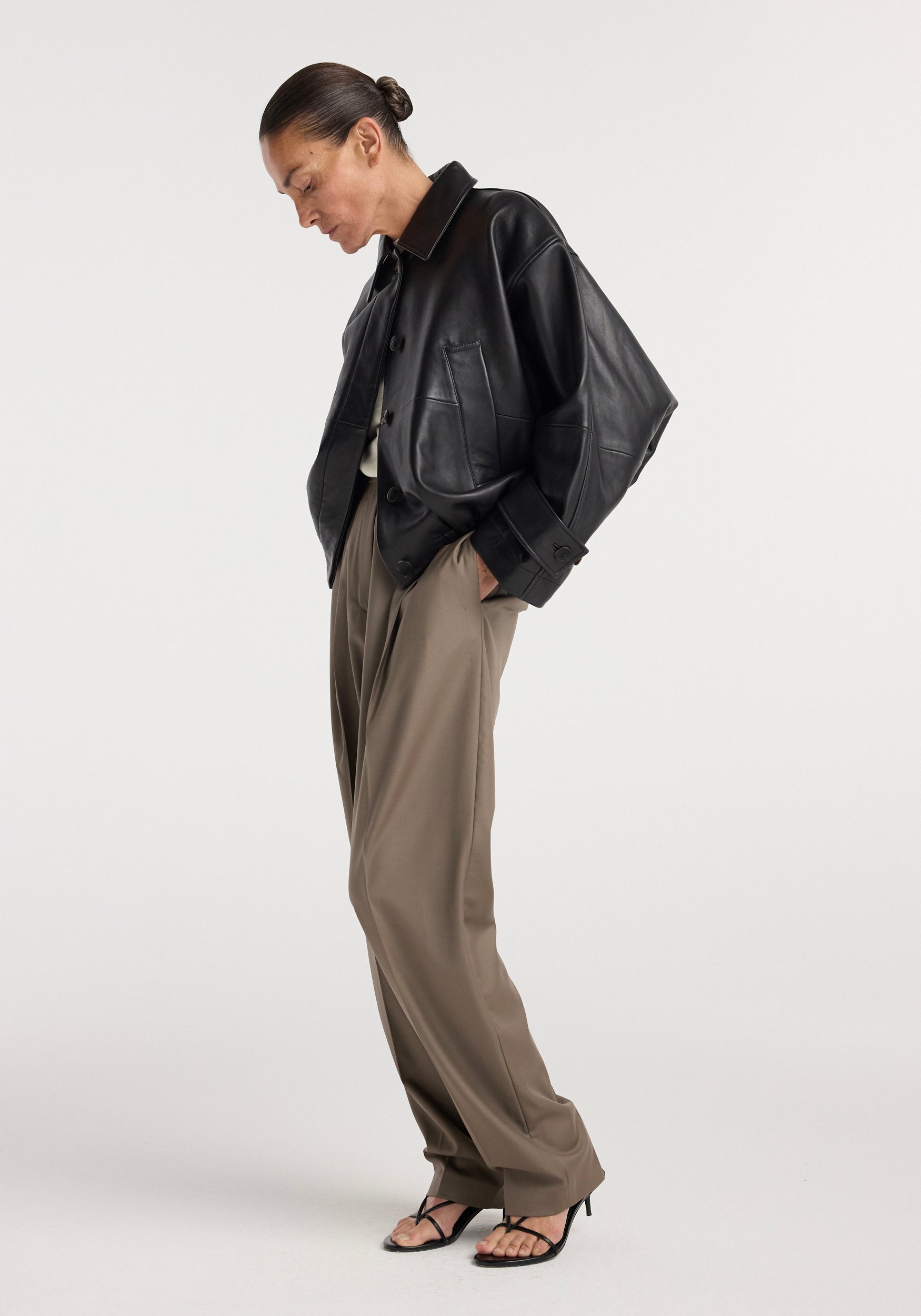 Woman wearing a black leather blouson jacket | black with oversized fit, paired with loose taupe pants and black strappy heeled sandals against a white background.