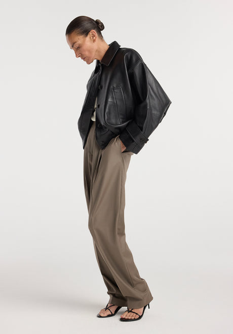 Woman wearing a black Leather blouson jacket | black with loose khaki pants and black strappy sandals, posing against a plain white background.