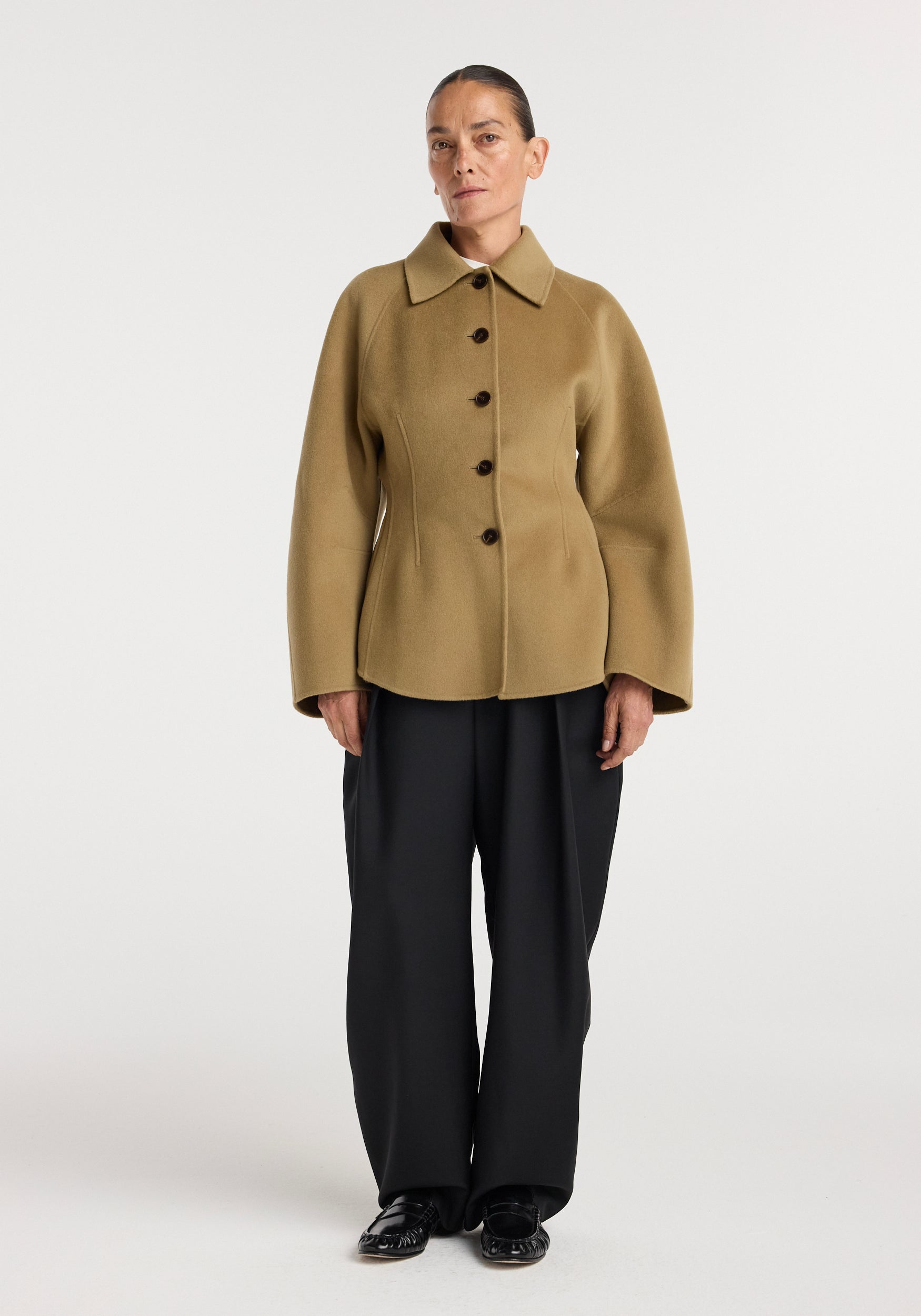 A model wearing a camel-colored double-faced sculptural jacket with a wide collar, button front, and voluminous sleeves paired with black wide-leg pants and black shoes.