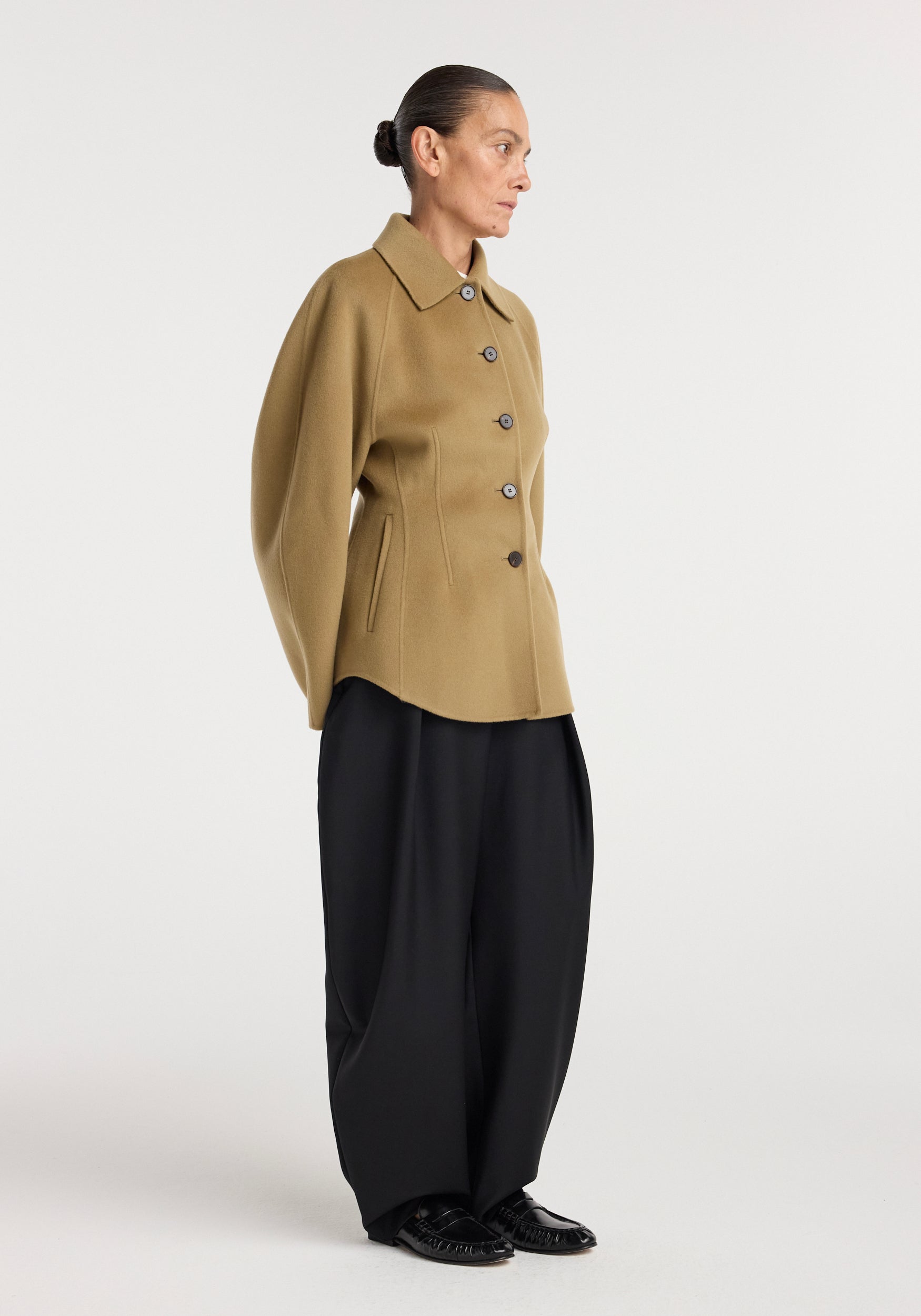 Model wearing a camel-colored Double-faced sculptural jacket | camel with button closure, structured shoulders, paired with loose black trousers and black shoes against a plain background.