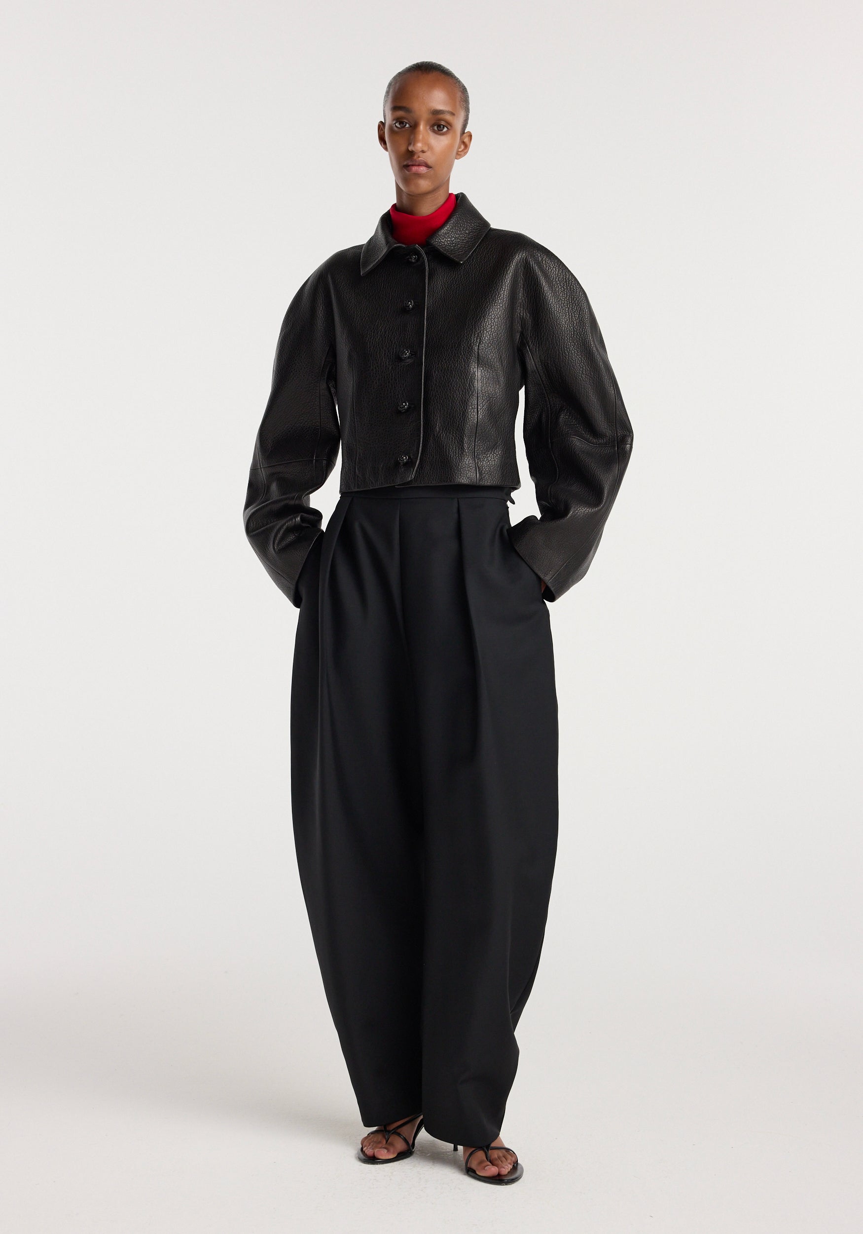 Model wearing a black Rounded grain leather cropped jacket | black with large button closure and voluminous sleeves, paired with high-waisted wide-leg black pants and black sandals.