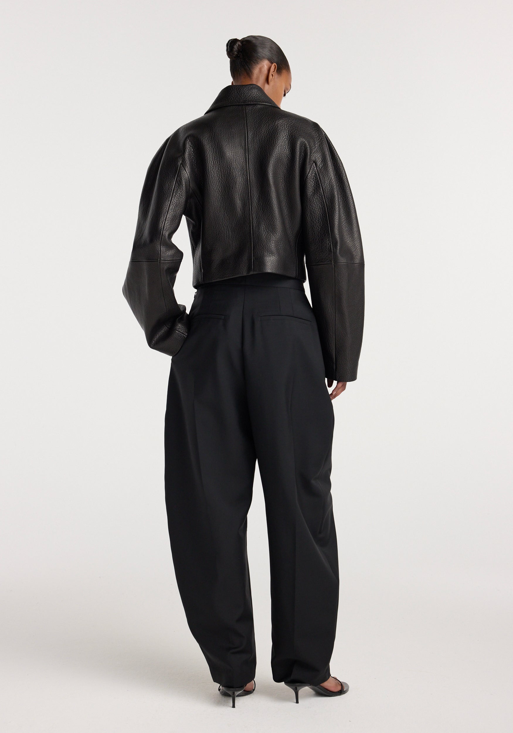 Back view of a woman wearing a black Rounded grain leather cropped jacket | black paired with loose black trousers and black open-toe heels against a plain background.