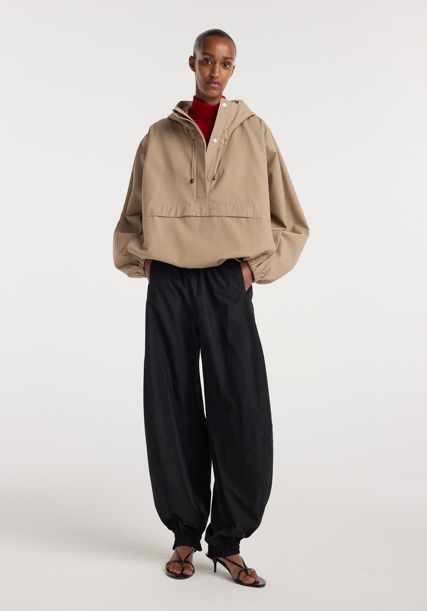 A model wearing a beige hooded anorak with a loose fit and long sleeves, paired with wide-leg black pants and open-toe heels, standing against a plain white background.
