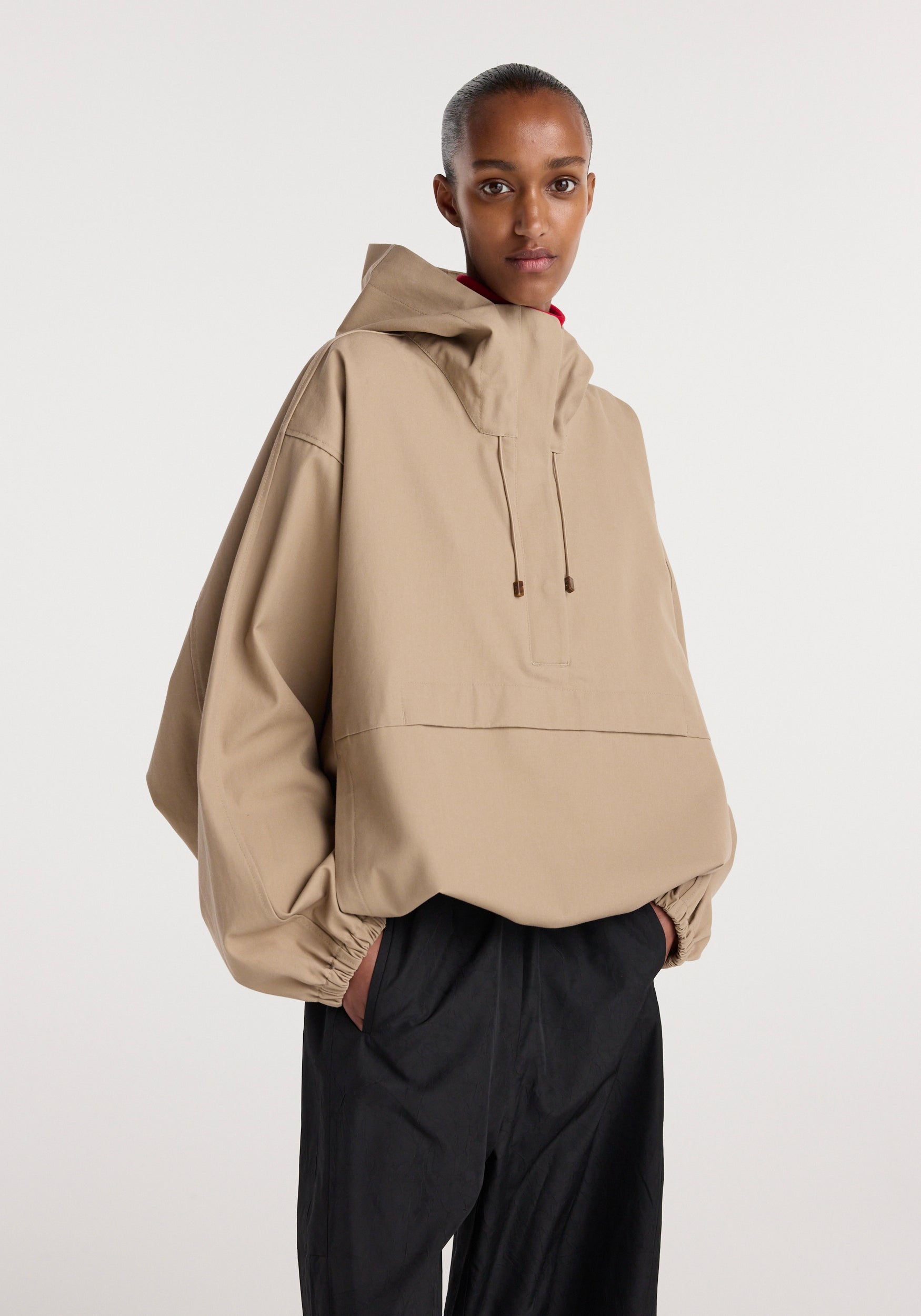 A model wearing a loose-fitting beige hooded anorak | beige with adjustable hood strings and elastic cuffs paired with black pants against a plain background.