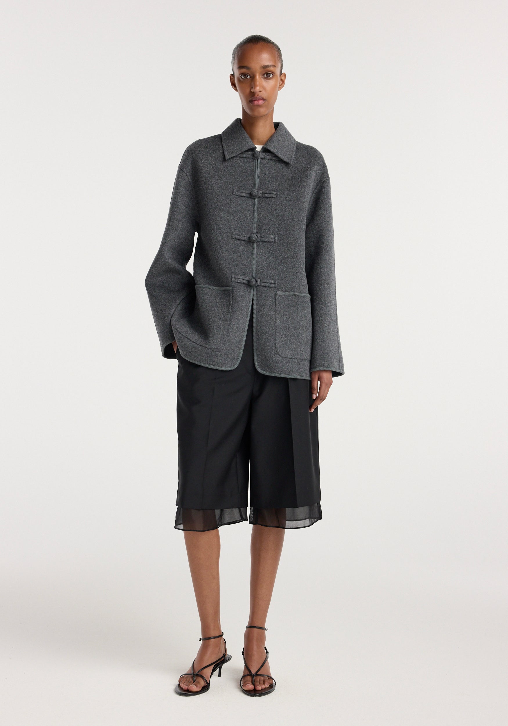 A model wearing a double-faced wool pankou closure jacket | dark grey melange, styled with black wide-leg shorts and strappy black heels against a neutral background.