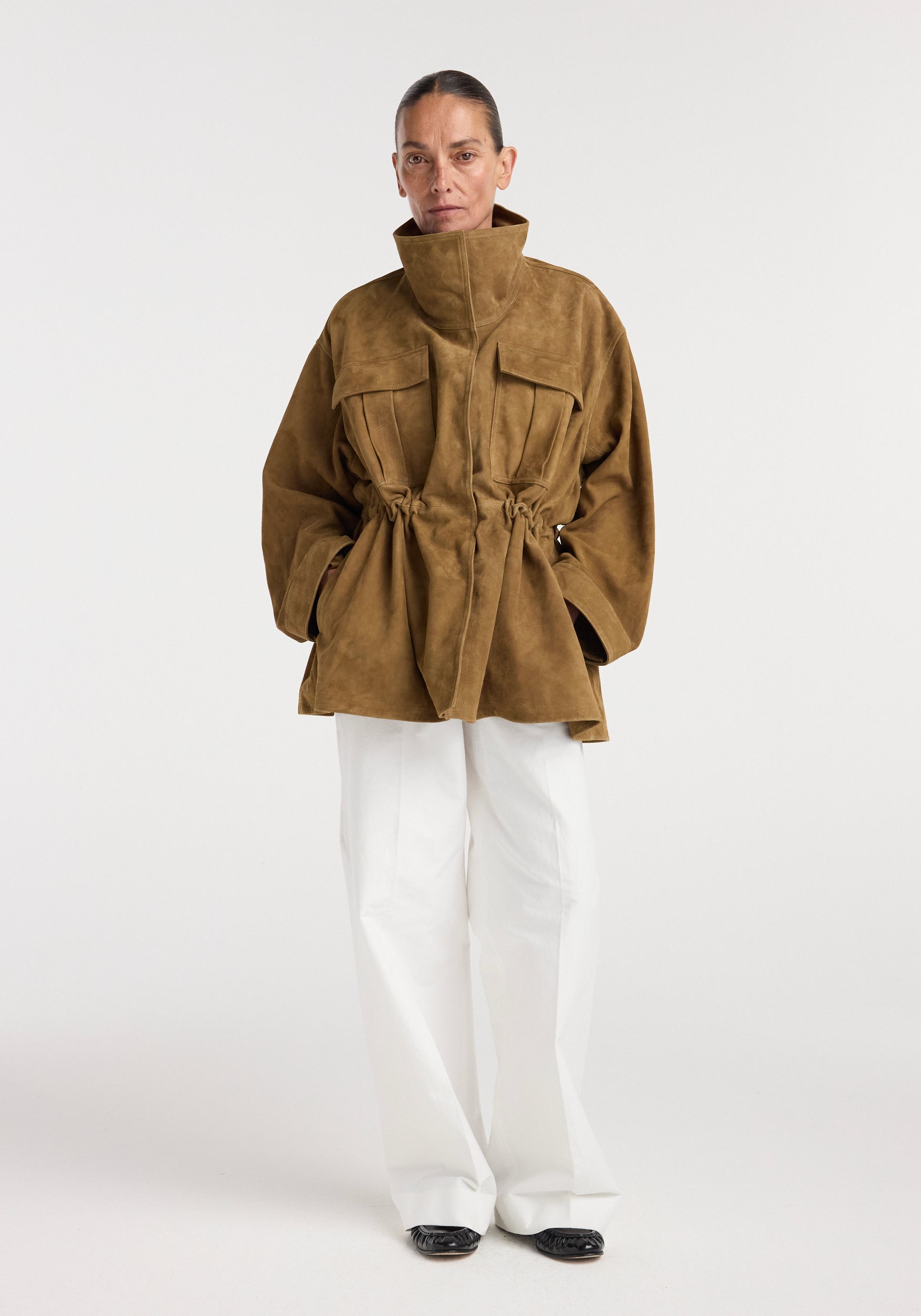 Woman wearing oversized sculptural suede jacket | sage in a brown tone with high collar and drawstring waist, paired with wide white pants and black shoes on plain background.
