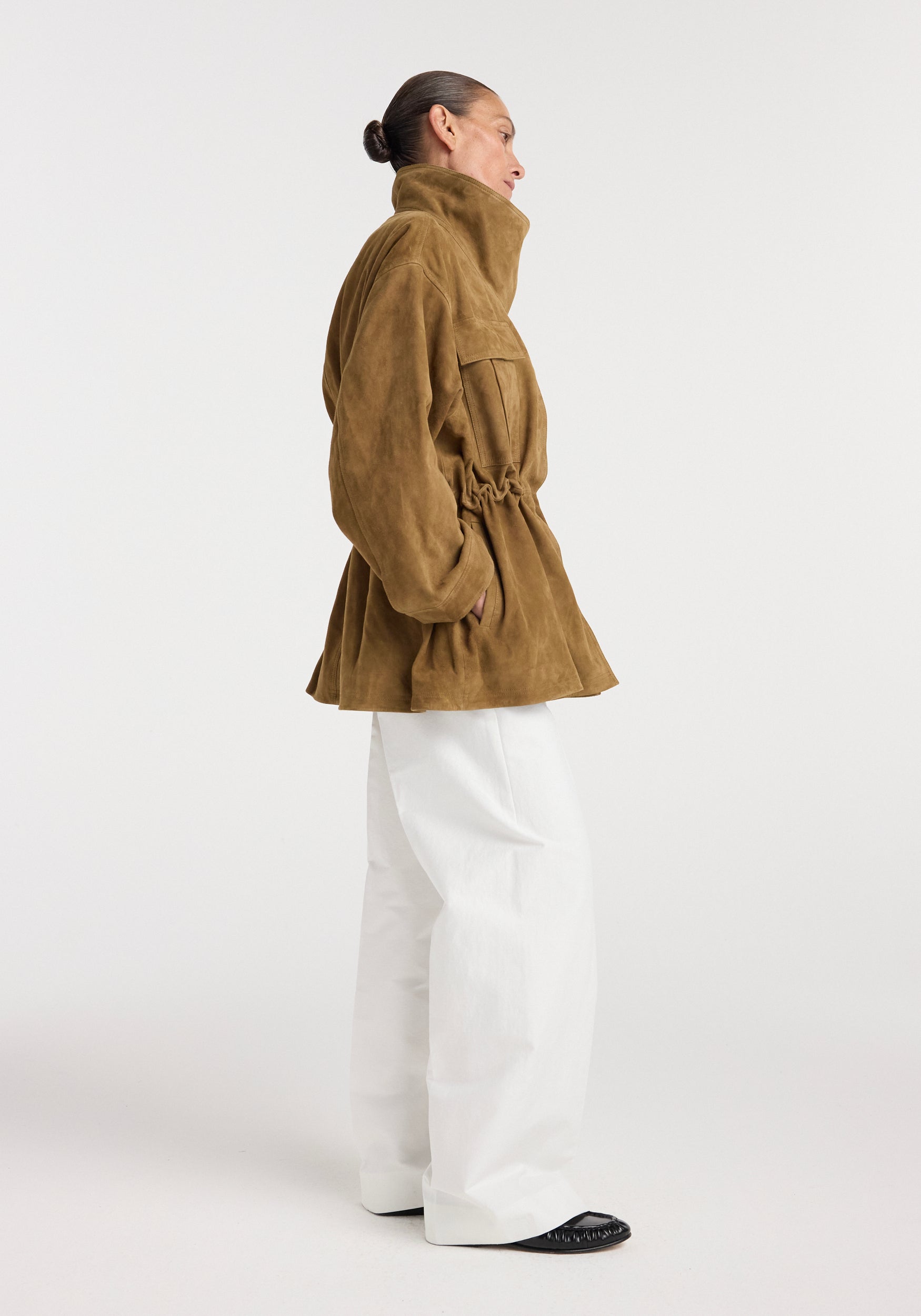 Side view of a woman wearing a sculptural suede jacket | sage in a relaxed fit with high collar, cinched waist, and pockets, paired with wide-leg white pants and black shoes.