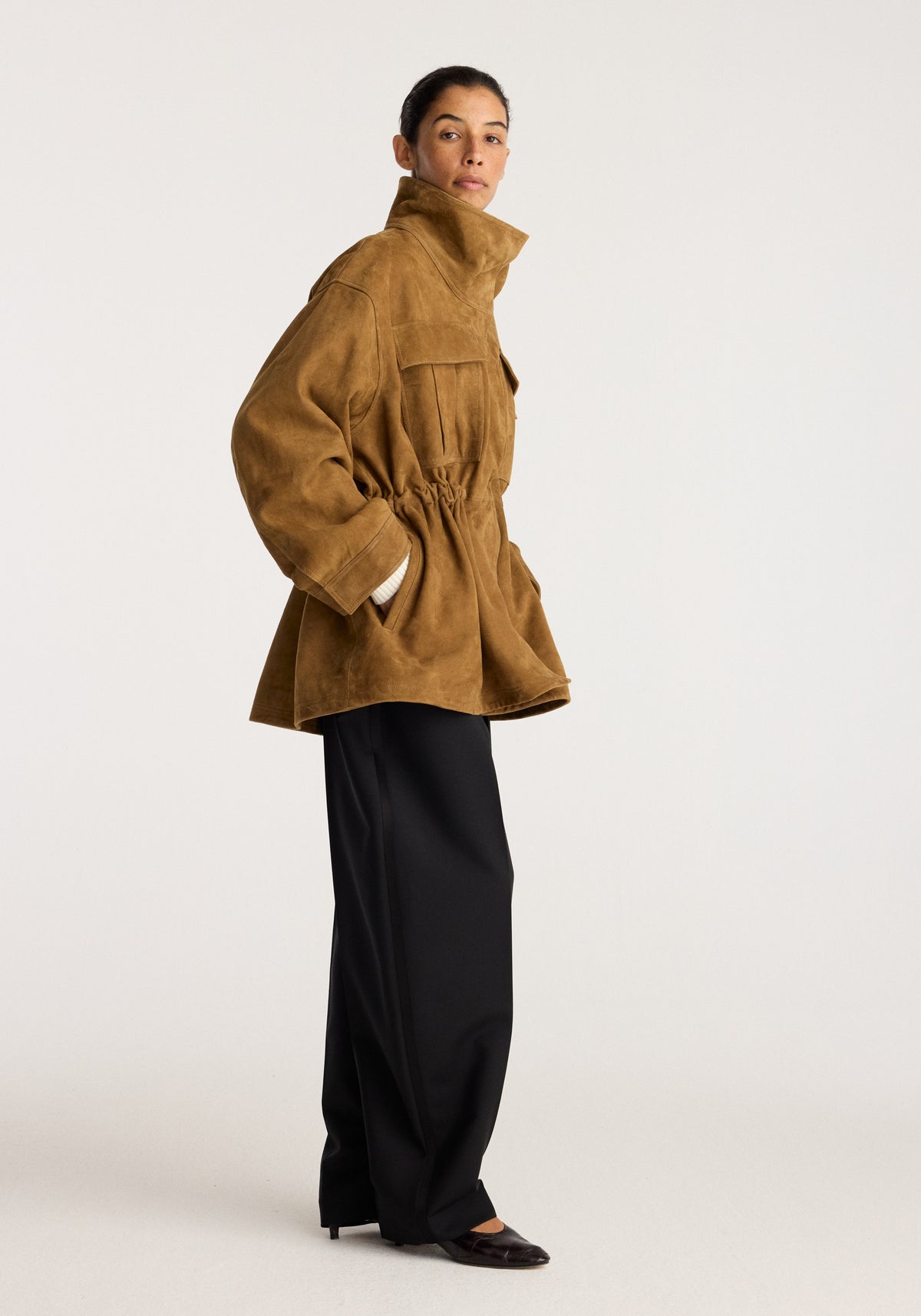 Sculptural suede jacket | sage worn by a model with a high collar gathered waist patch pockets and flared hem styled with black wide leg trousers